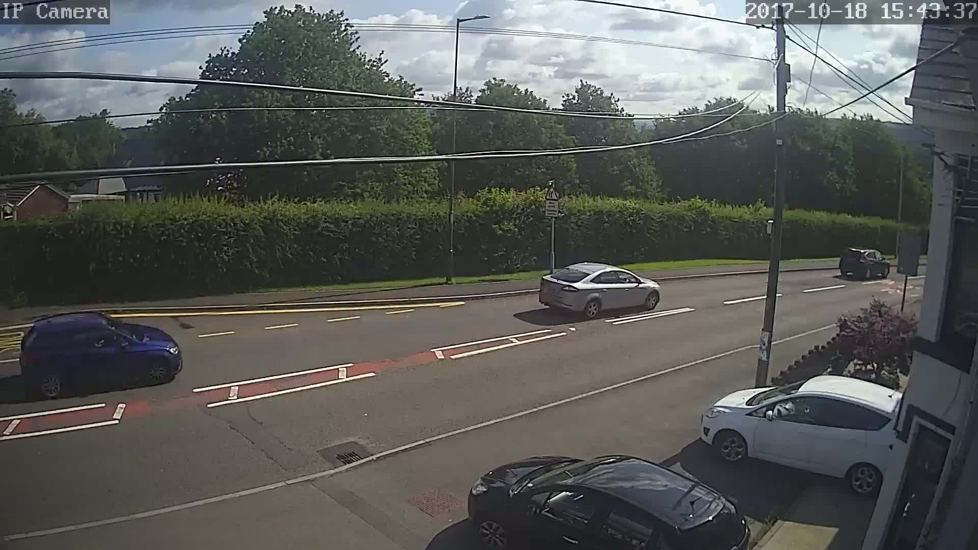 A sunny day shows several cars driving on a paved road lined with hedges and trees, with one car parked on the side.