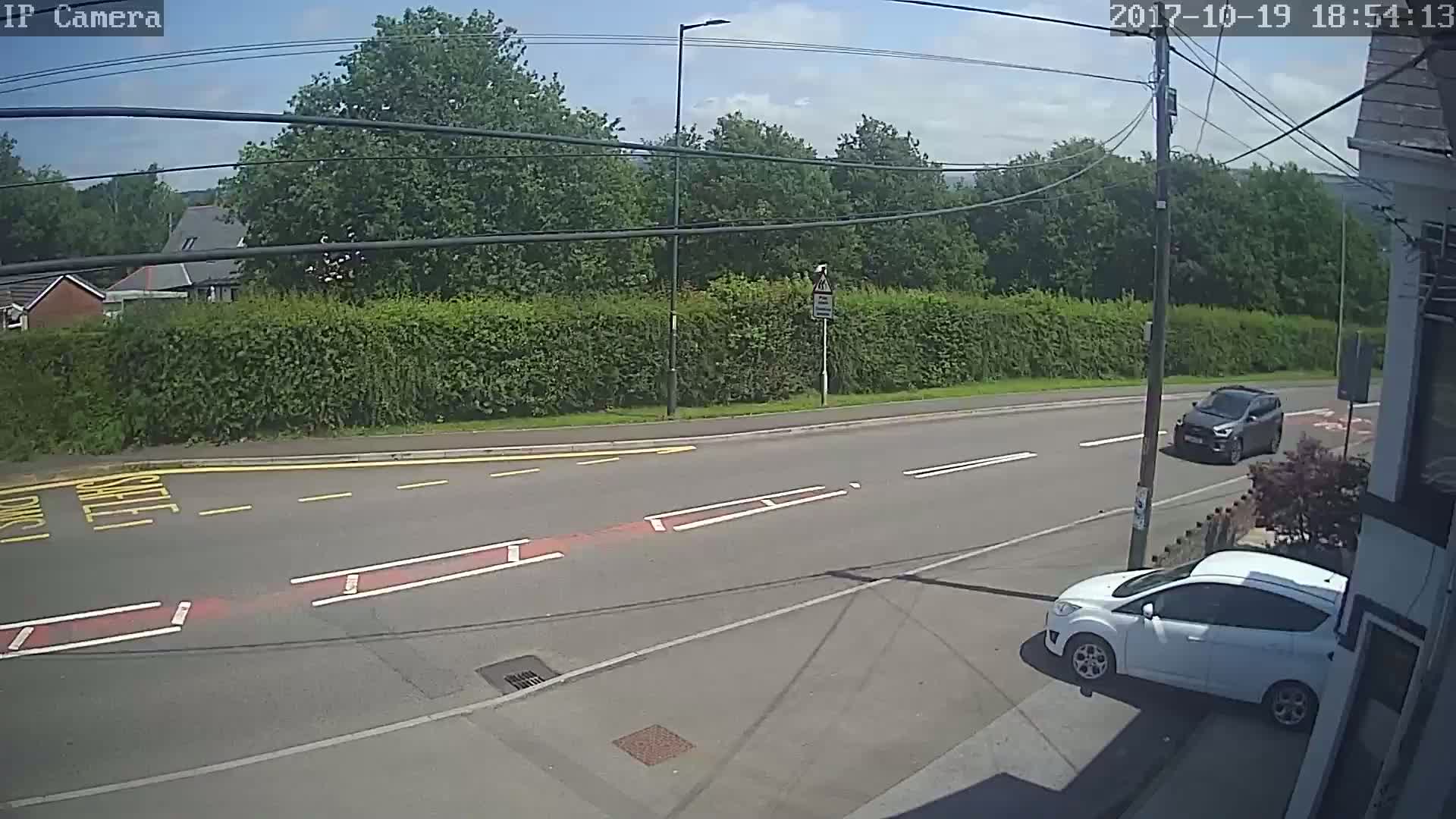 A sunny day shows a street scene with two cars, one dark-grey and one white, parked near a tall hedge bordering a road with yellow and red markings.
