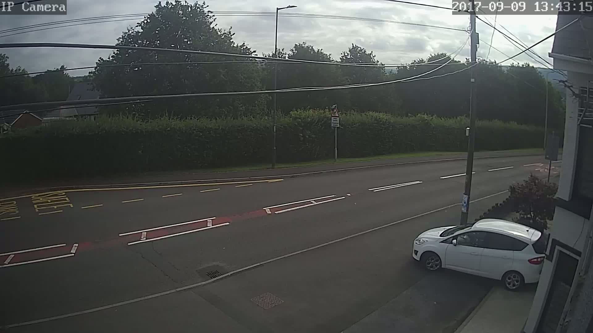 A white car is parked on the side of a road, which is bordered by a tall hedge and trees under a partly cloudy sky.