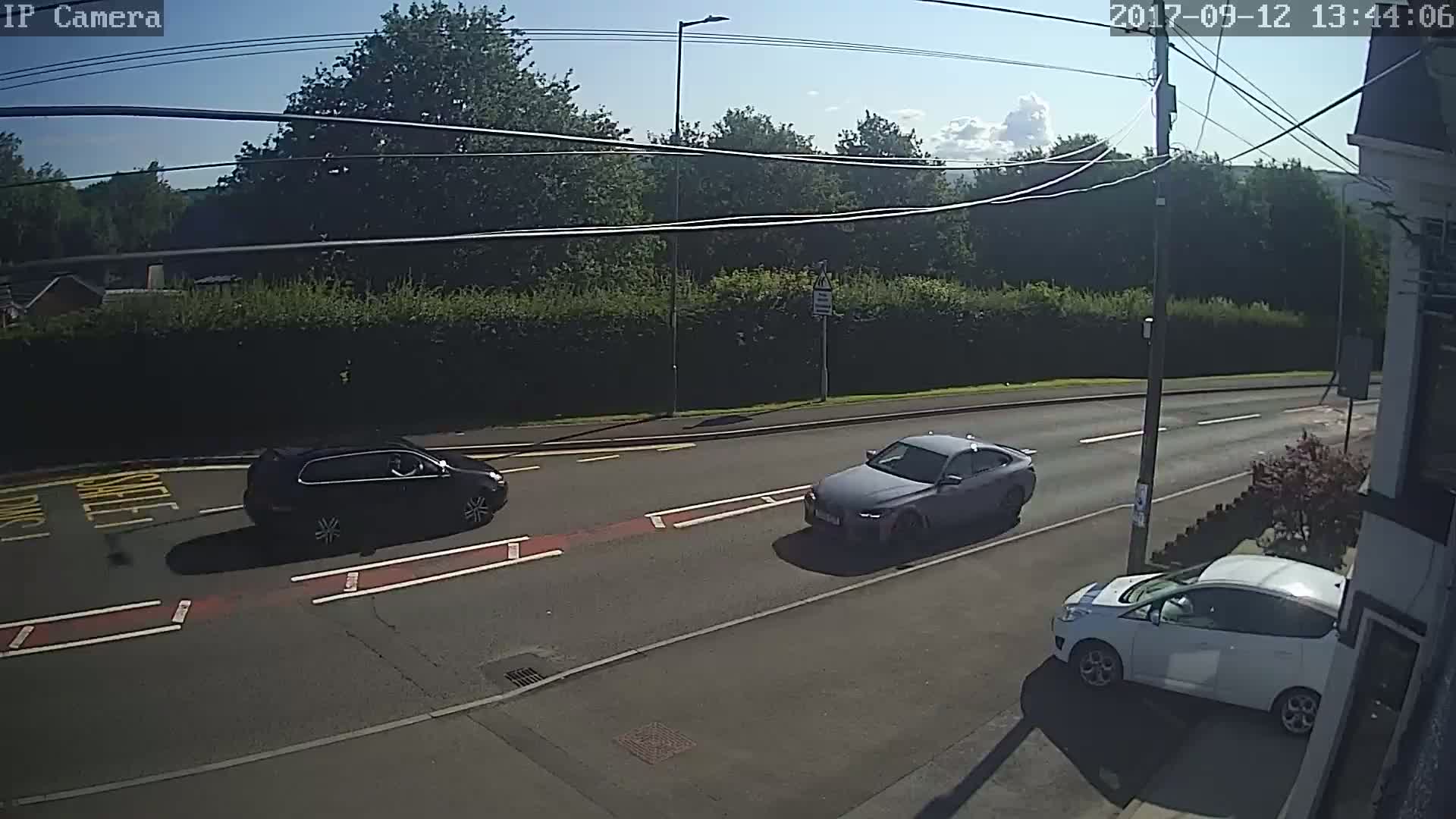 Three cars are visible on a sunny road lined with hedges and trees.