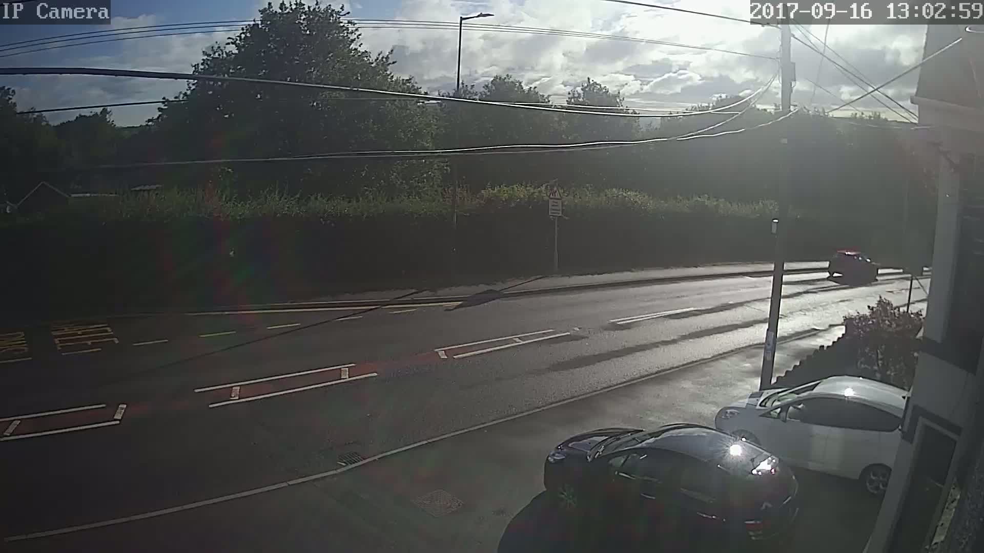 A partly sunny day shows a wet road with two cars parked near a building and one driving away from the camera, all set against a backdrop of greenery and power lines.
