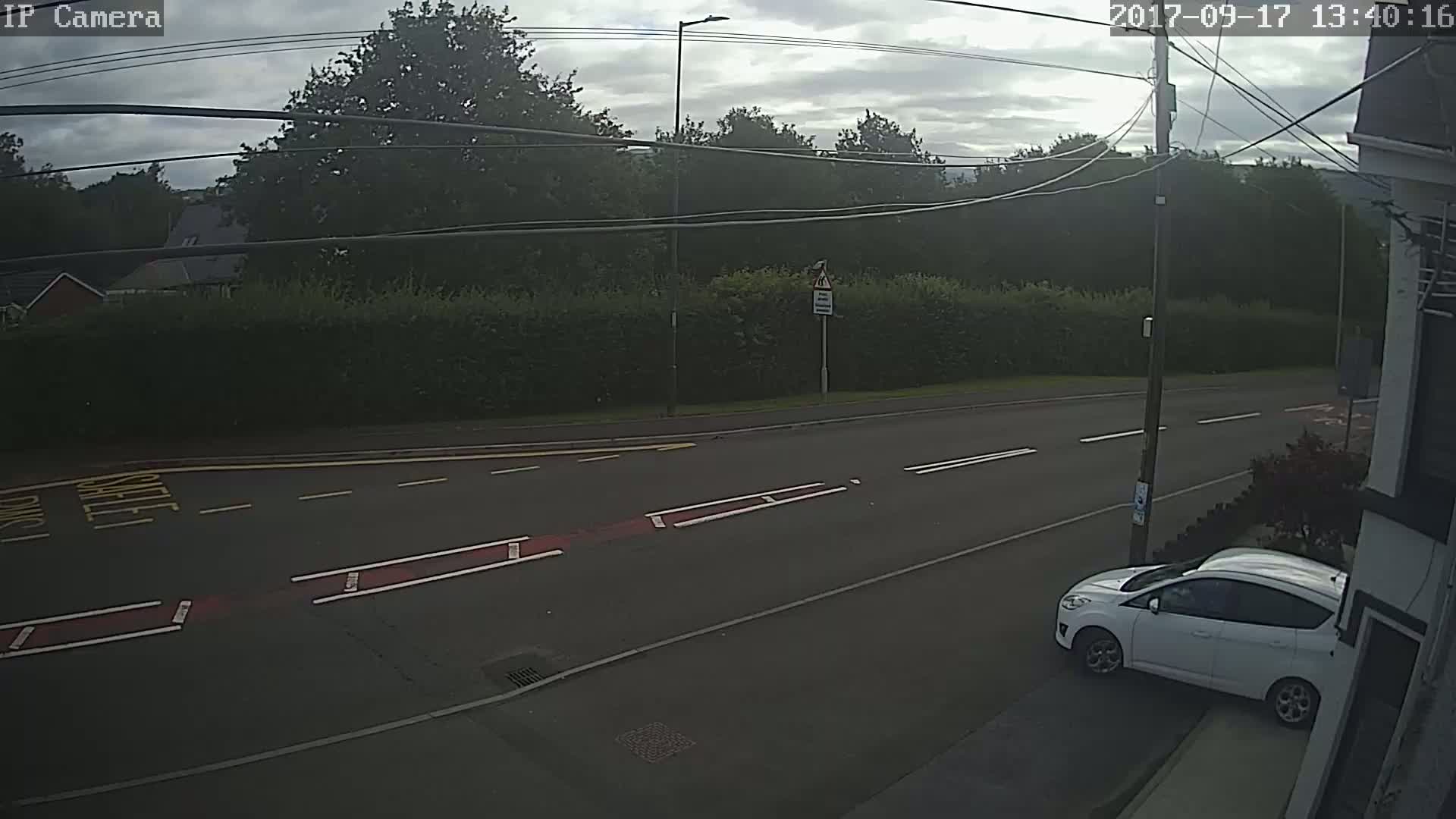 A white car is parked on the side of a road under an overcast sky, near a tall hedge and several power lines.