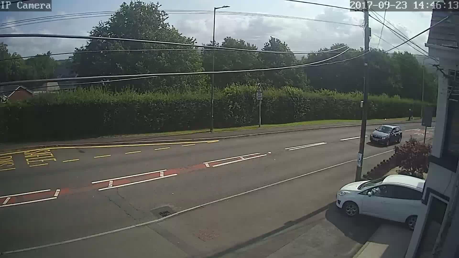 A sunny day shows a road with two cars, one parked and one driving,  and a hedge separating the road from a line of trees.