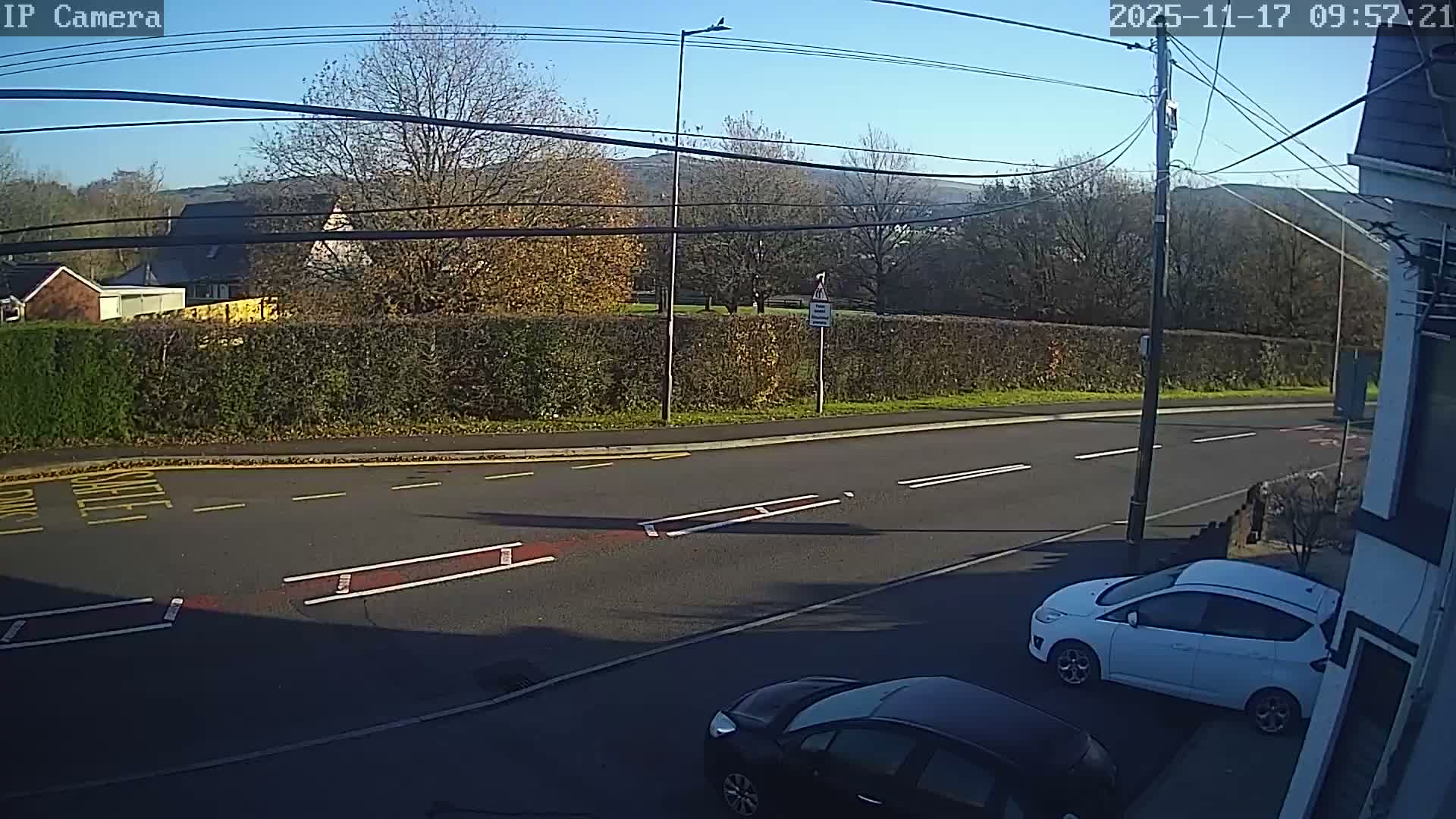 A clear and sunny day illuminates a residential street with parked cars, a hedge-lined road, and trees displaying autumn foliage, with distant hills visible under a bright blue sky.