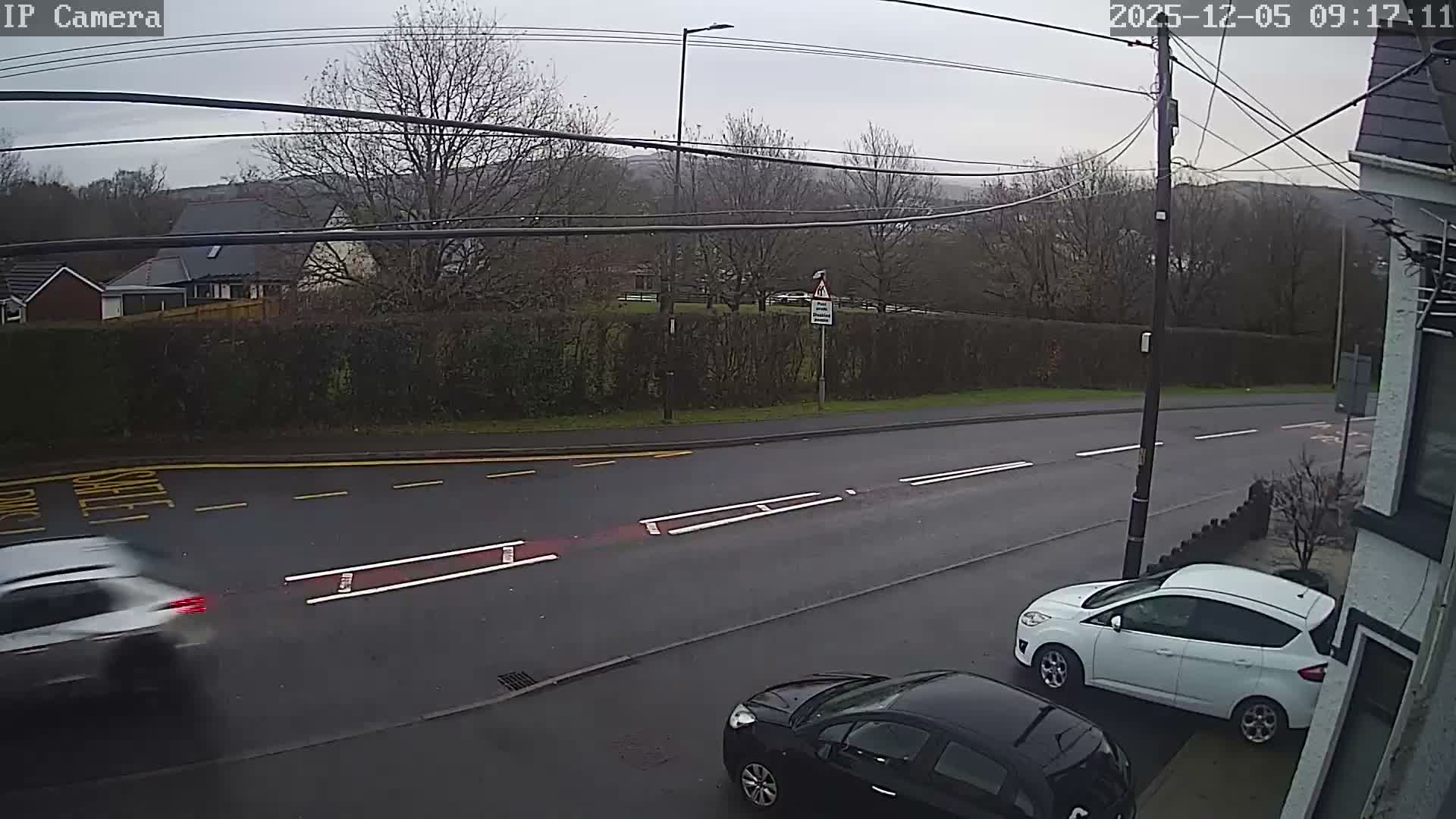 The image displays an overcast day on a residential street with a damp road, featuring a white car partially parked on the pavement, a black car parked behind it, a blurry silver car in motion on the road, and houses and bare trees lining the background.