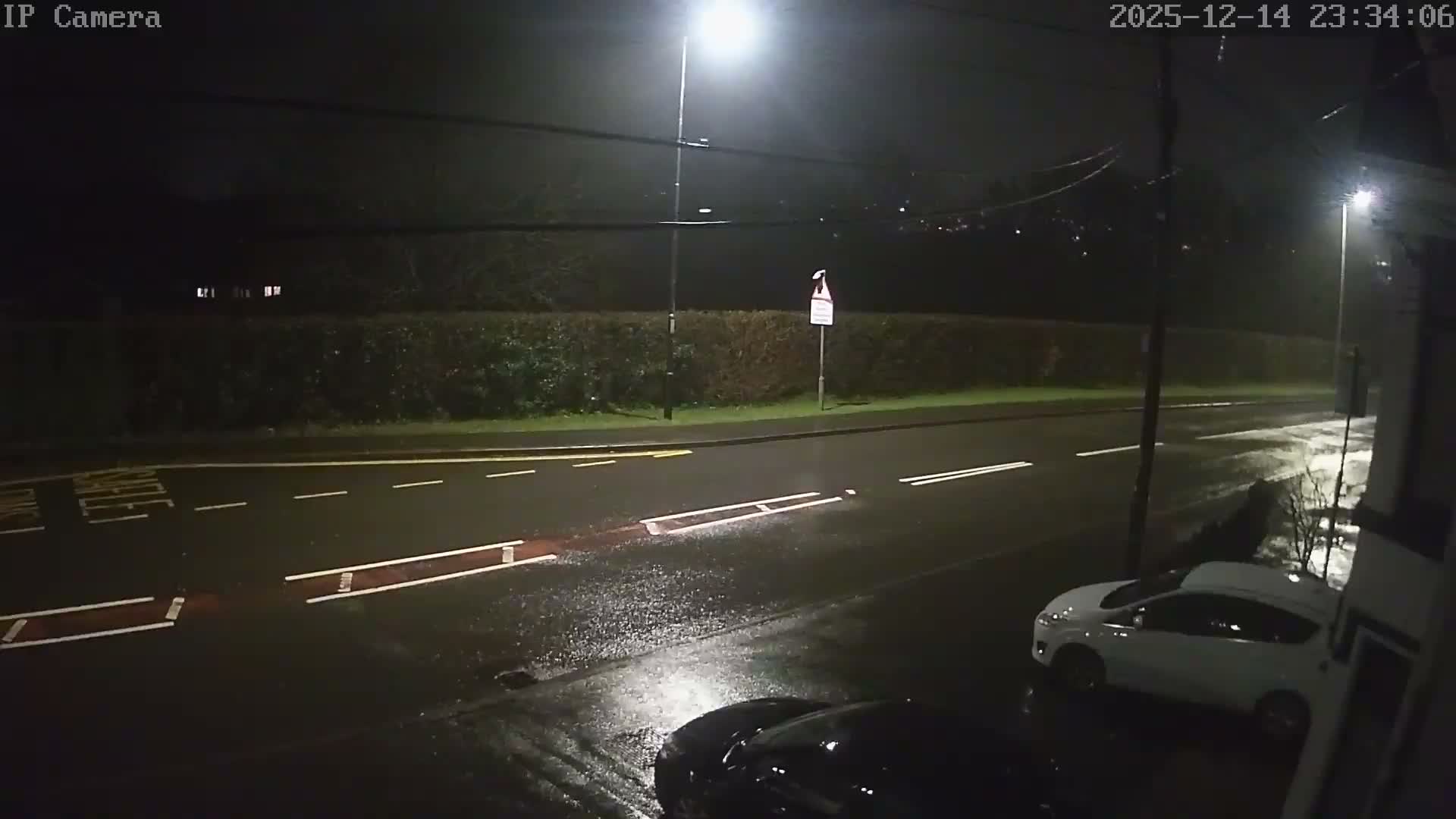 The image displays an overcast day on a residential street with a damp road, featuring a white car partially parked on the pavement, a black car parked behind it, a blurry silver car in motion on the road, and houses and bare trees lining the background.