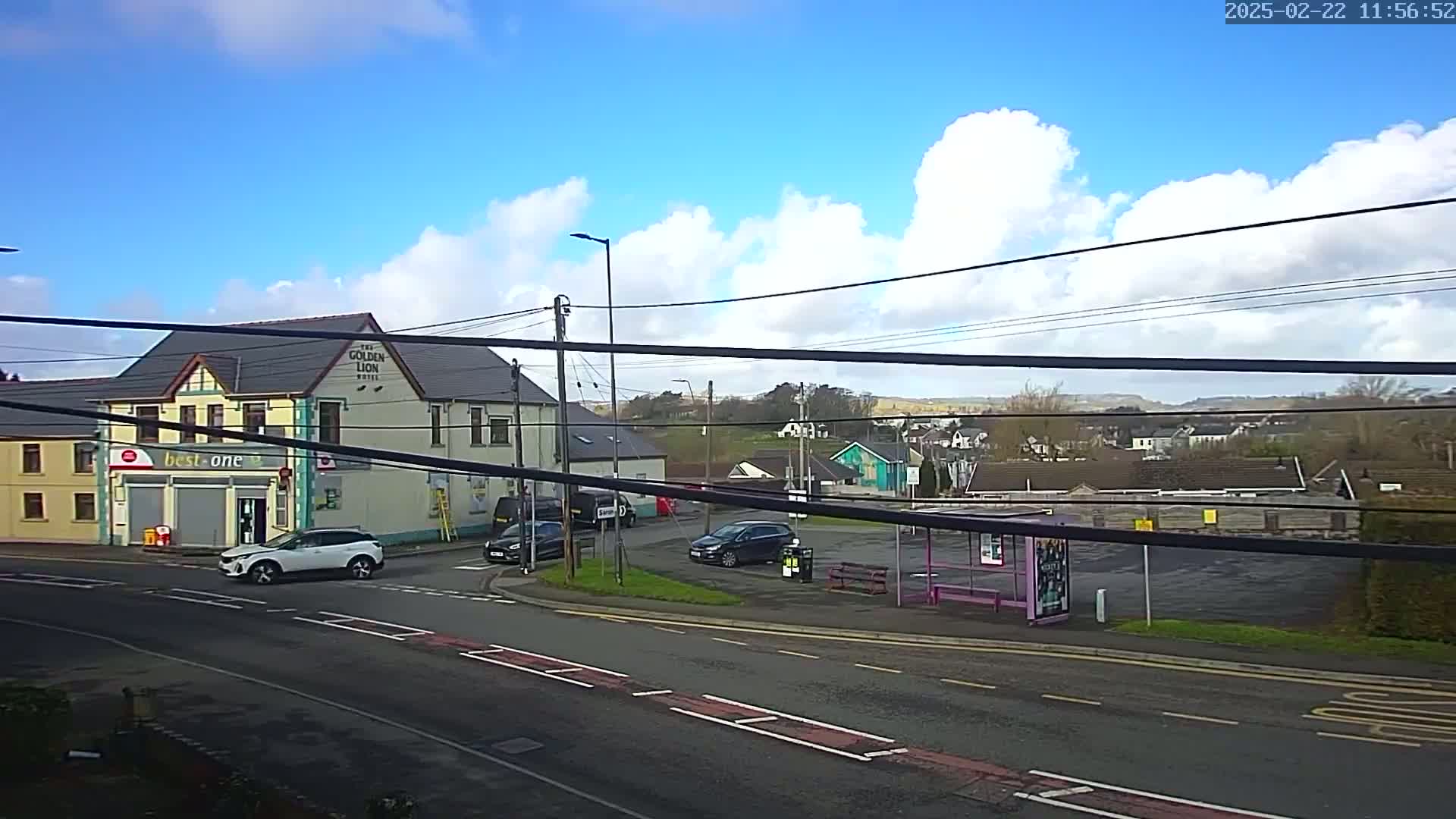 A mostly sunny day reveals a street scene with a two-story building, a few cars, and a bus stop, all situated near a hillside.