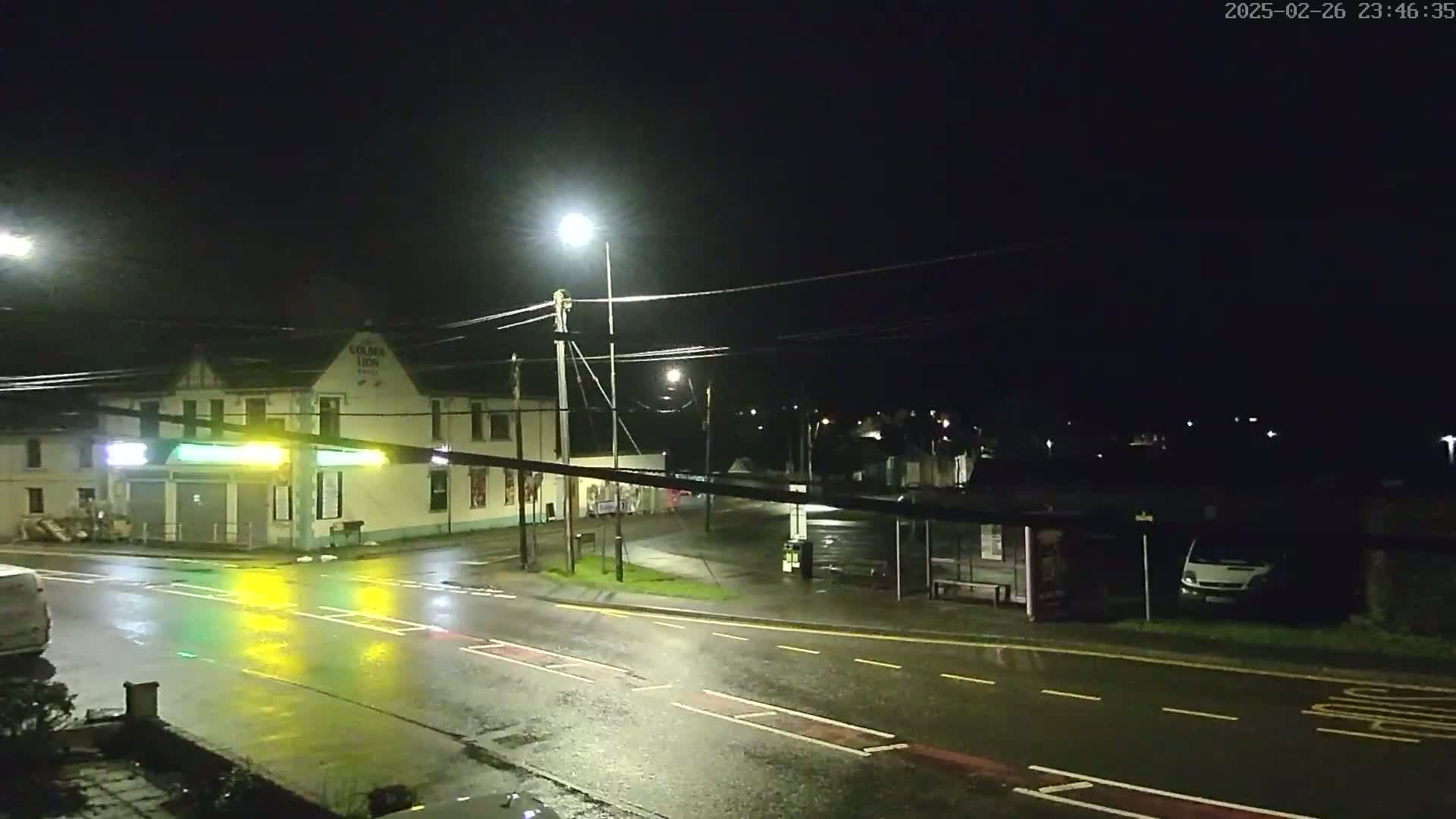 A wet, nighttime street scene shows a building with lights, a bus stop, and a van parked nearby, under several streetlights.
