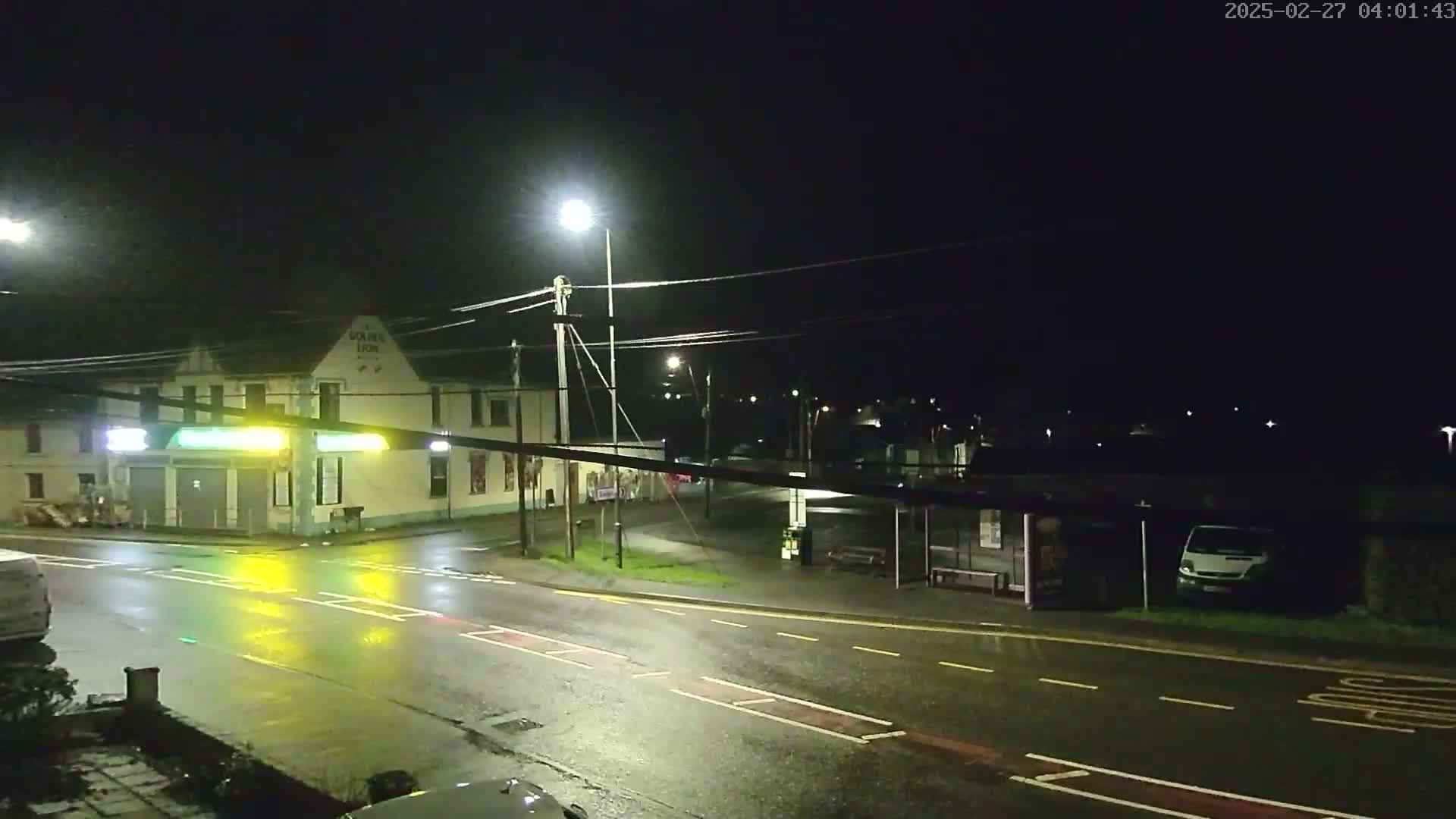 A wet, nighttime street scene shows a building with lights on, a bus shelter, and a few vehicles.