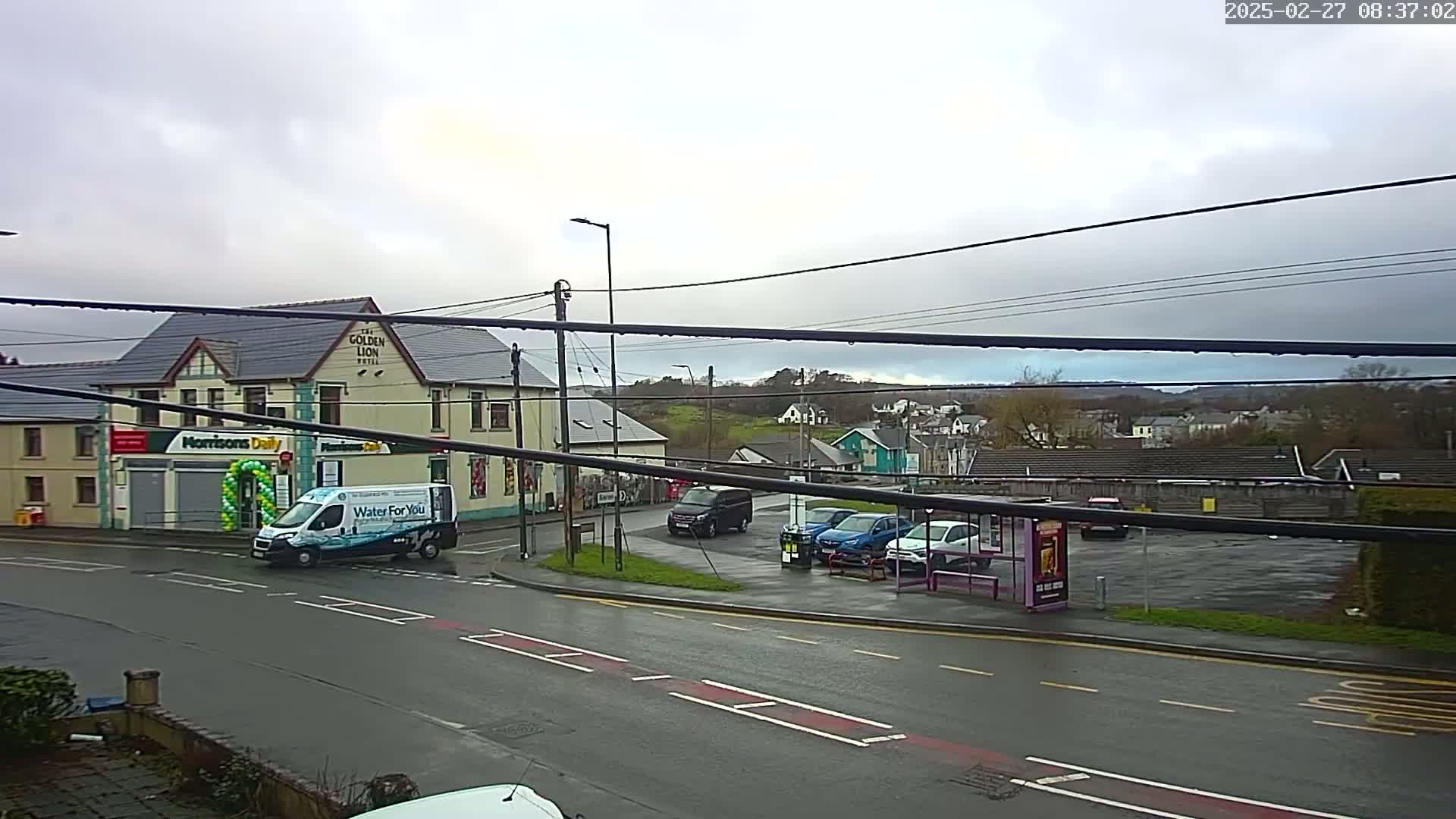 A wet, overcast day reveals a street scene with parked cars, a small van, a corner store, and a pub in the background, along with a small hill in the distance.