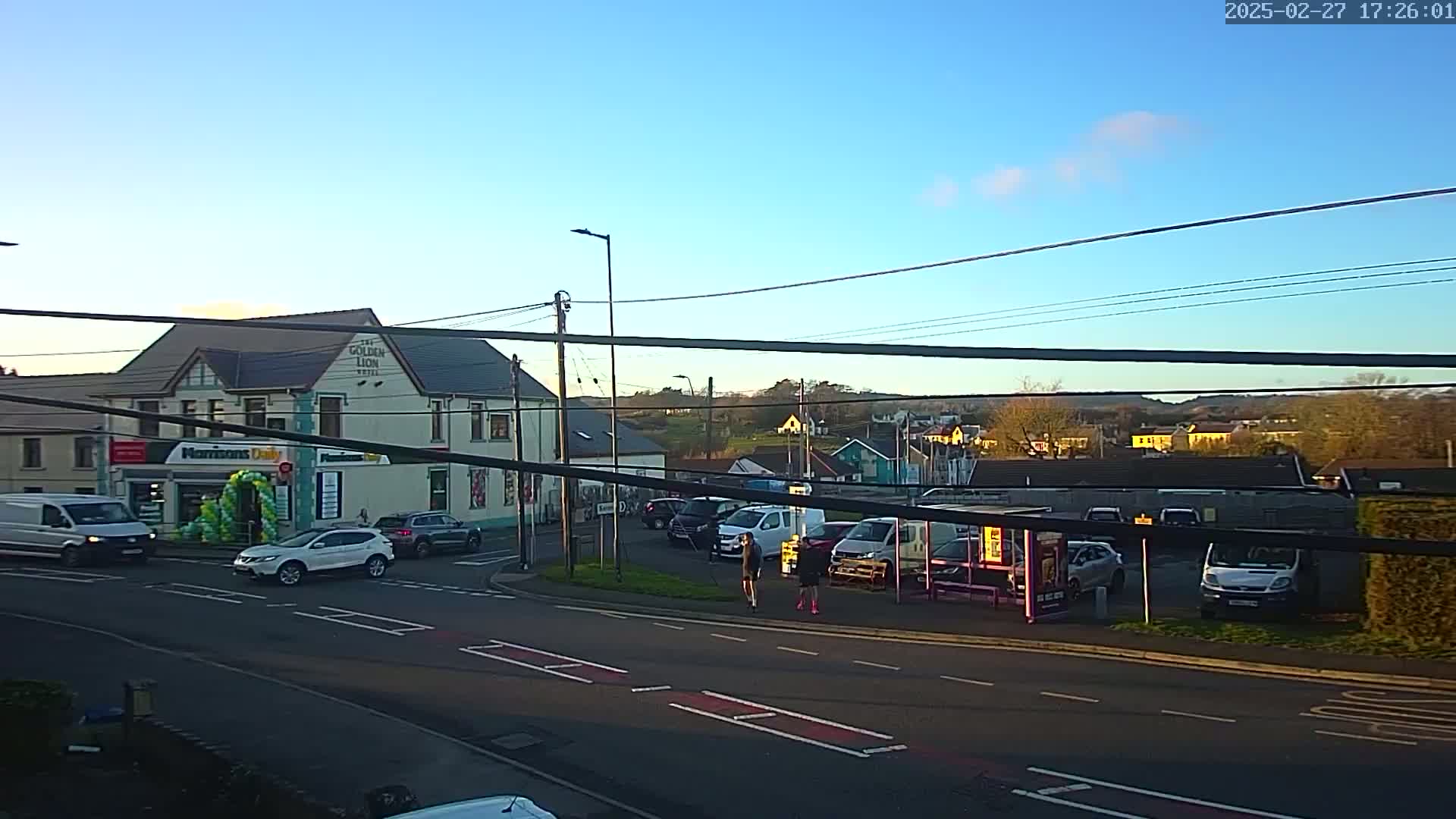 A street scene on a sunny day shows cars parked and driving near a pub and a small store, with two pedestrians crossing the road.