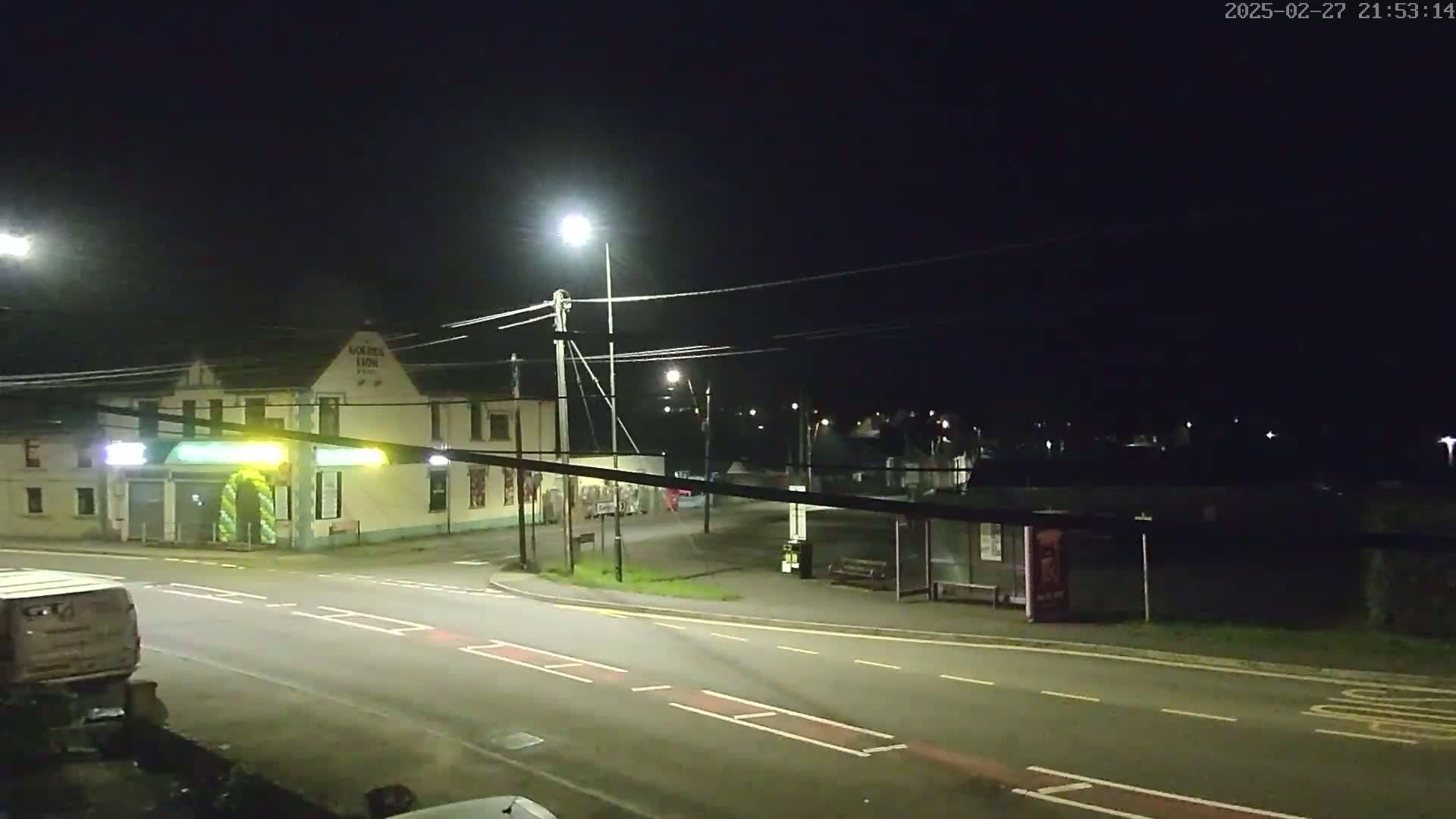 A nighttime street scene shows a mostly empty road curving past a pub and bus stop, illuminated by streetlights under a clear, dark sky.