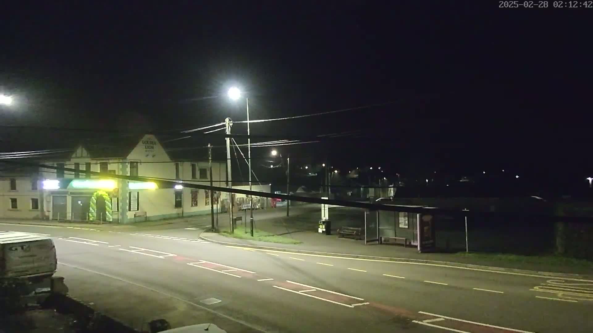 A nighttime street scene shows a building with illuminated signage, a bus stop, and a vehicle partially visible on the left, under a clear, dark sky.
