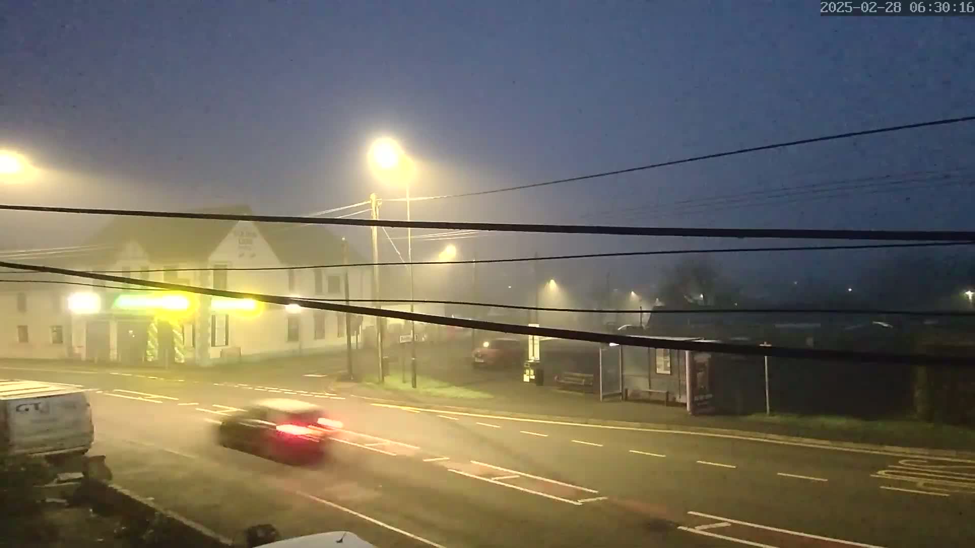A foggy intersection at night, with several cars driving on the road past a building and bus shelter illuminated by streetlights.