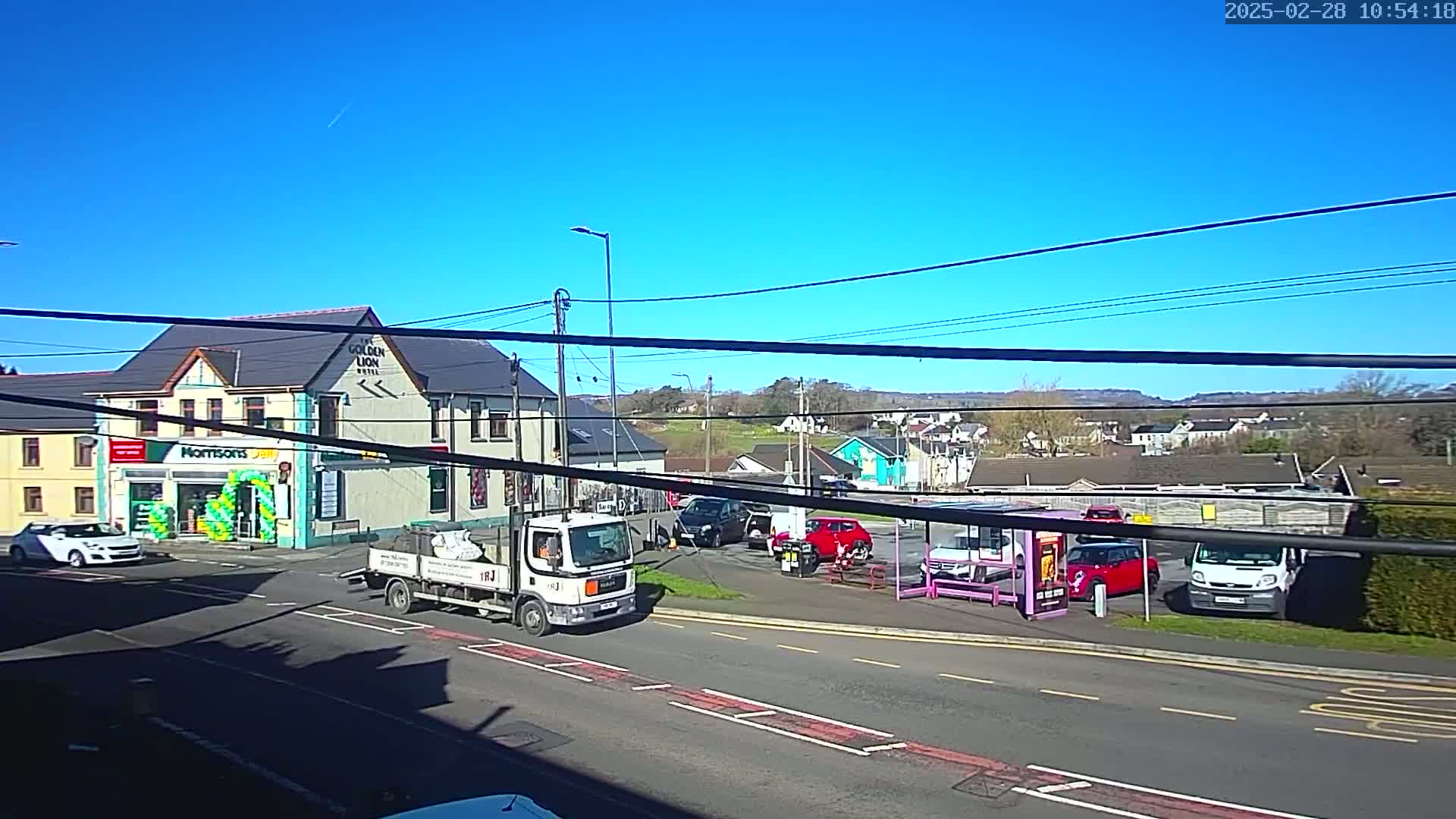 A sunny day shows a street scene with several cars and a delivery truck parked near a pub and a convenience store, all situated in a small town with distant hills.