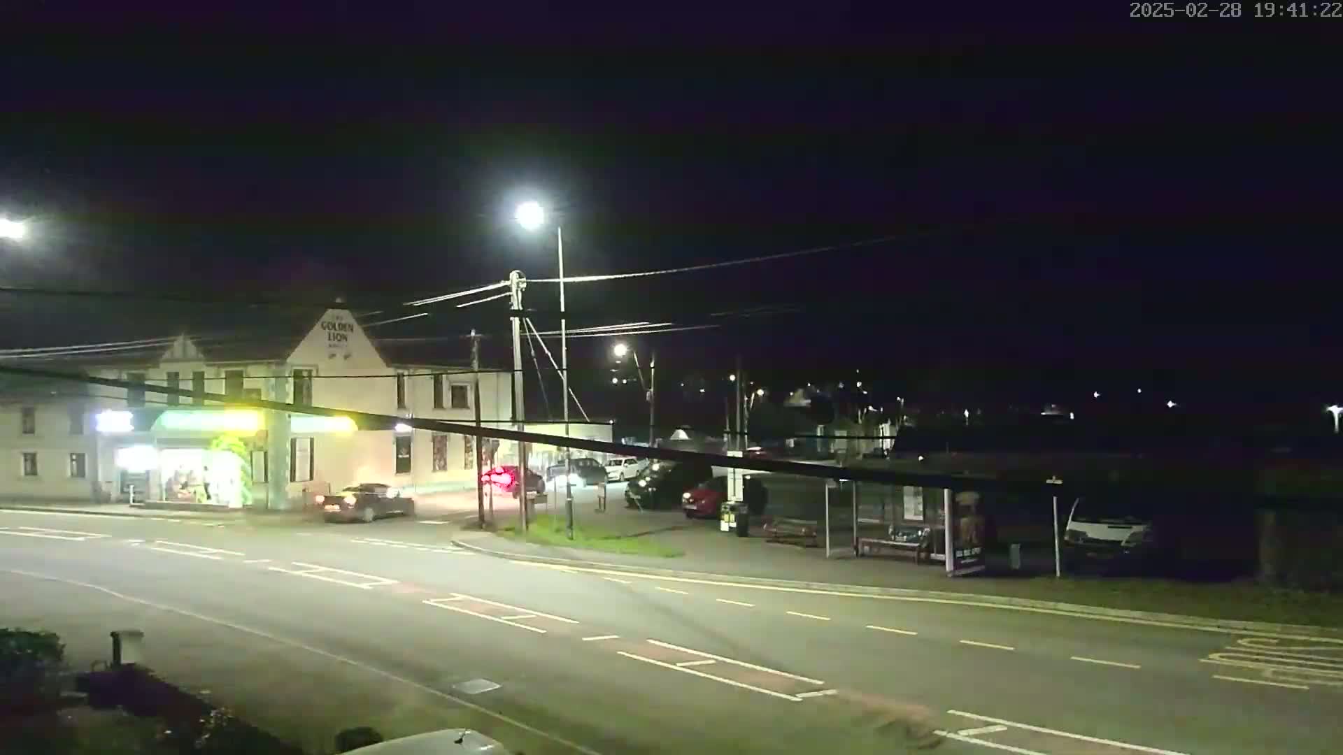 A nighttime street scene shows a road intersection with several cars, a bus stop, and a building lit by streetlights.