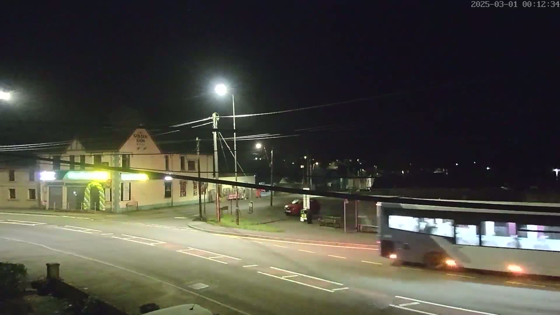 A nighttime street scene shows a bus passing a pub and a car parked near a bus stop under streetlights.
