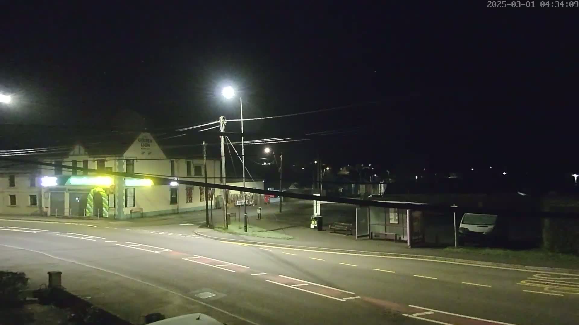 A nighttime street scene shows a road intersection with a pub, a bus stop, and a van parked nearby, under dark, clear skies illuminated by streetlights.