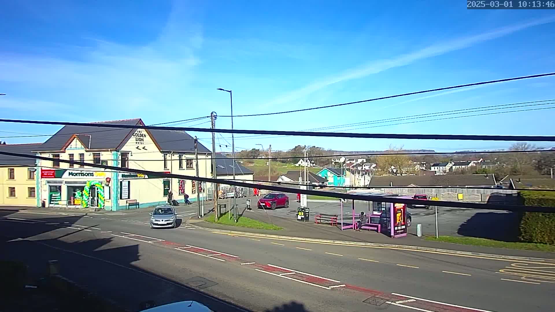 A sunny day reveals a street scene with a two-story building, a small shop, several cars, a bus stop, and a residential area in the background.