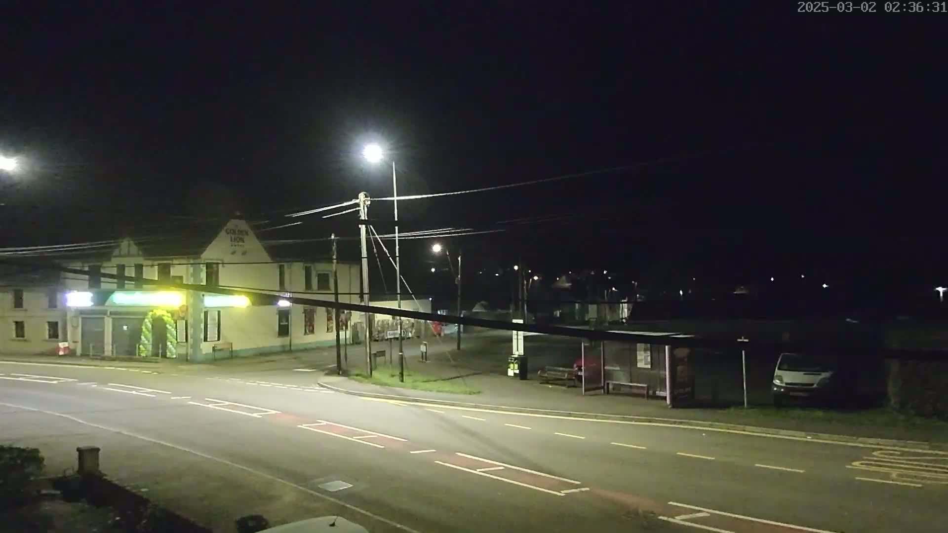A nighttime street scene shows a light-colored building, a bus stop, a van, and several streetlights illuminating a roadway intersection under a clear, dark sky.