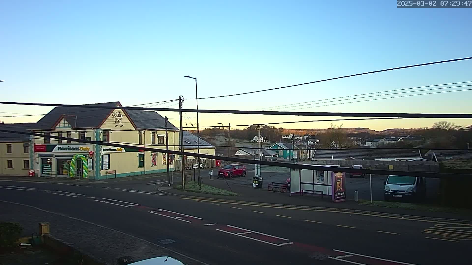 A clear sunrise illuminates a town square with a yellow building, a small convenience store, a bus shelter, and several parked cars.