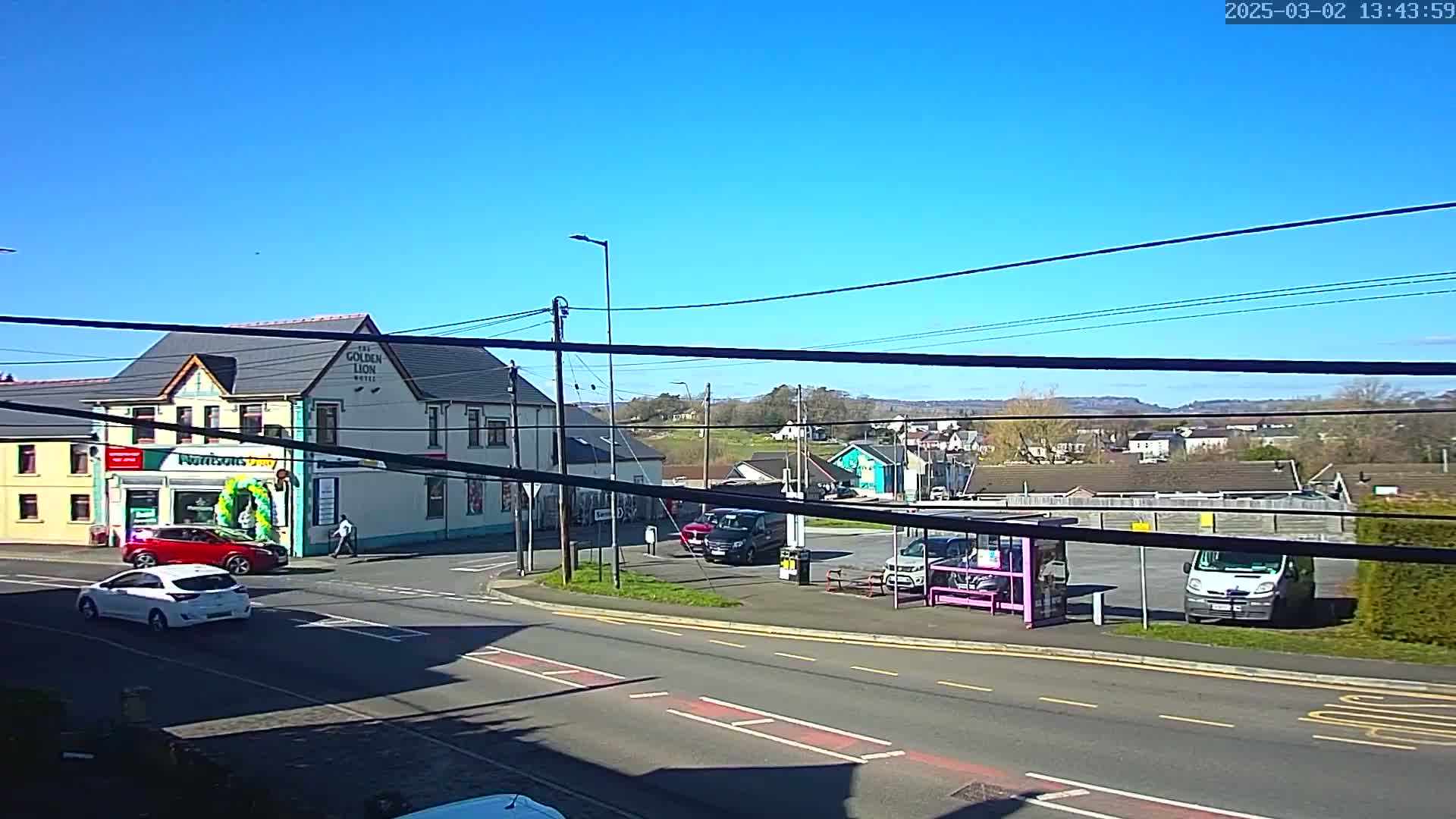 A sunny day shows a town square with cars parked near a building, a bus stop, and other buildings in the background.
