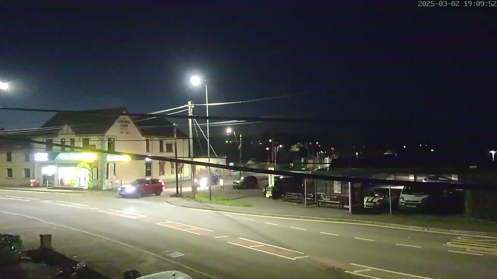 A nighttime street scene shows a two-story building with a few cars, a bus shelter, and some parked vehicles under streetlights.
