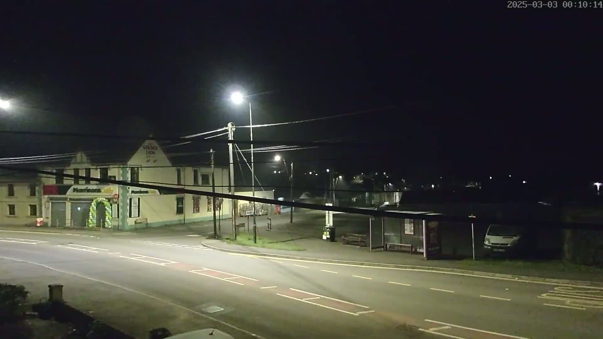 A nighttime street scene shows a road intersection with a building, a bus shelter, and a van parked nearby under artificial light.
