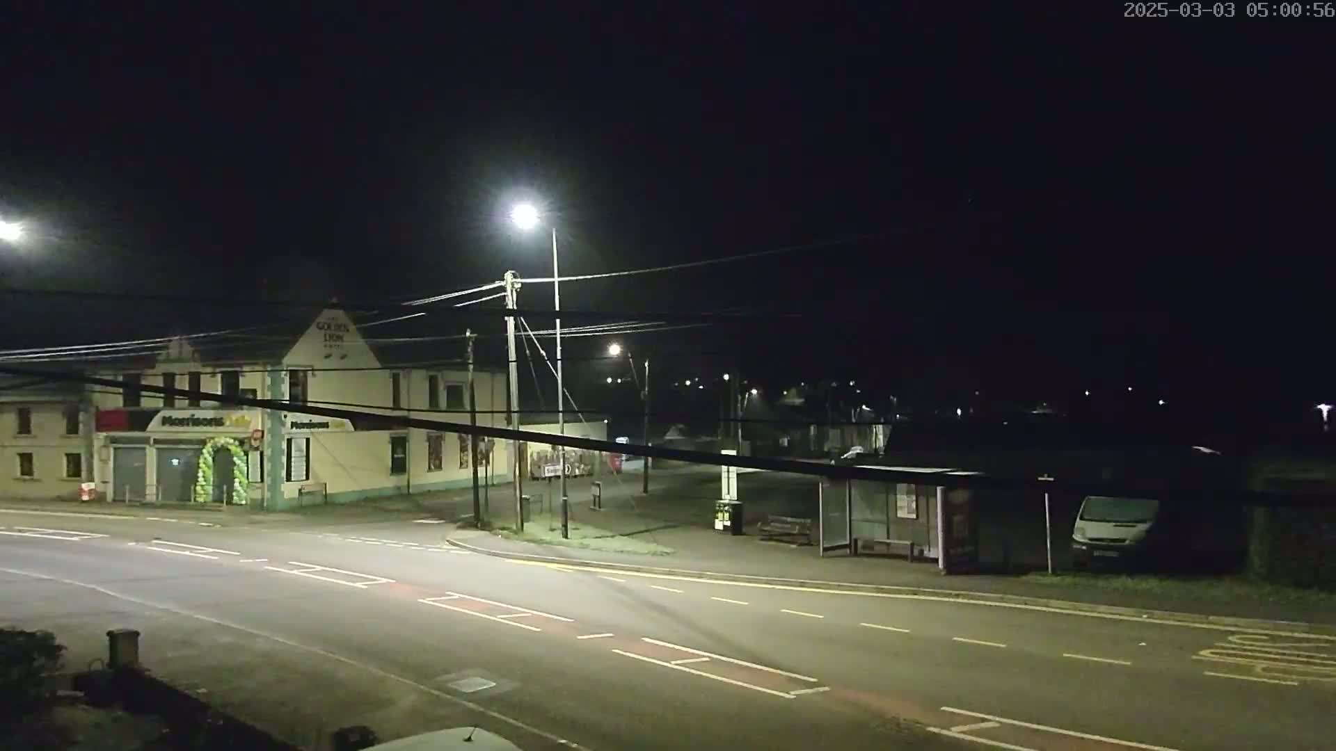 A nighttime street scene shows a building with lights on, a bus stop, and a van parked nearby, under dark skies.