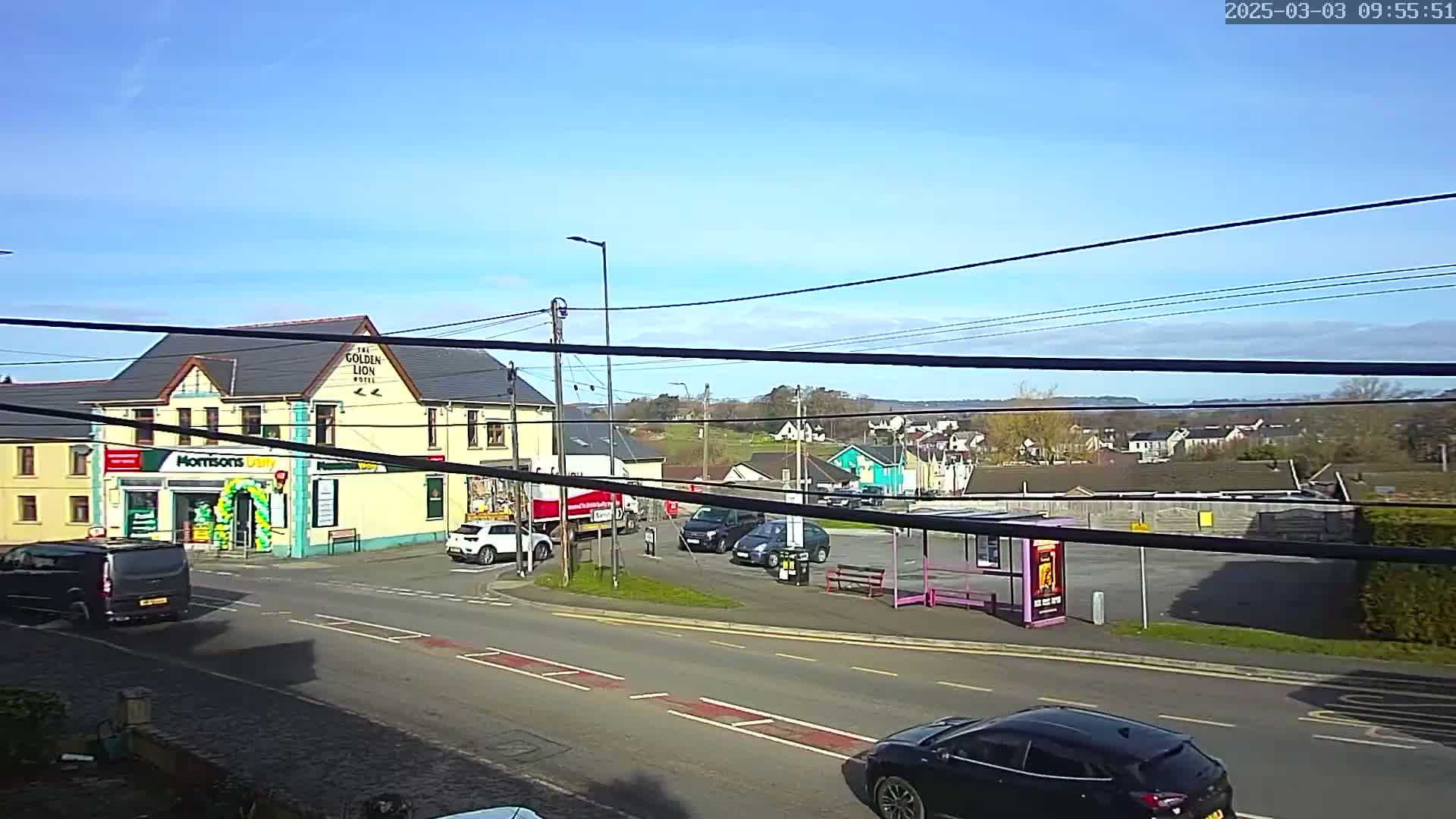 A sunny day reveals a street scene with a two-story building housing a store and hotel, several cars, a bus stop, and a distant residential area.