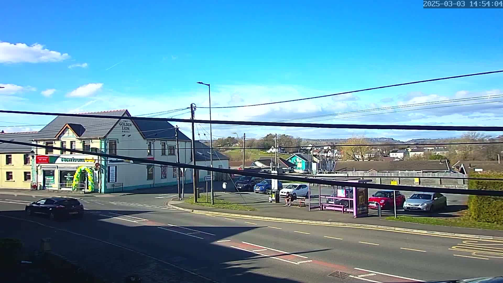 A sunny day reveals a street scene with a two-story building, several cars parked near a bus stop, and a distant view of houses and hills.