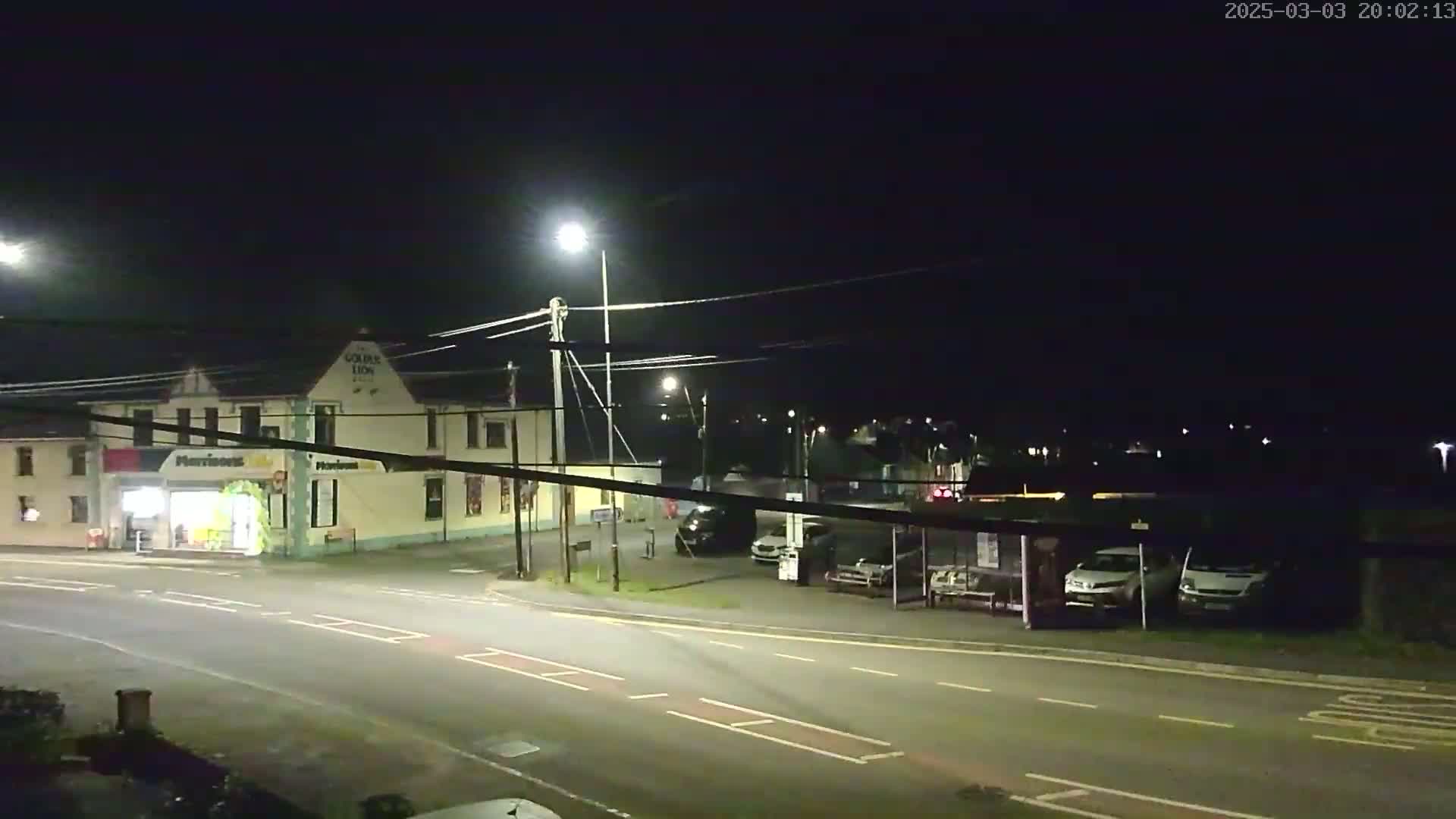 A nighttime street scene shows a building with a storefront, several parked cars, and a bus stop under streetlights.