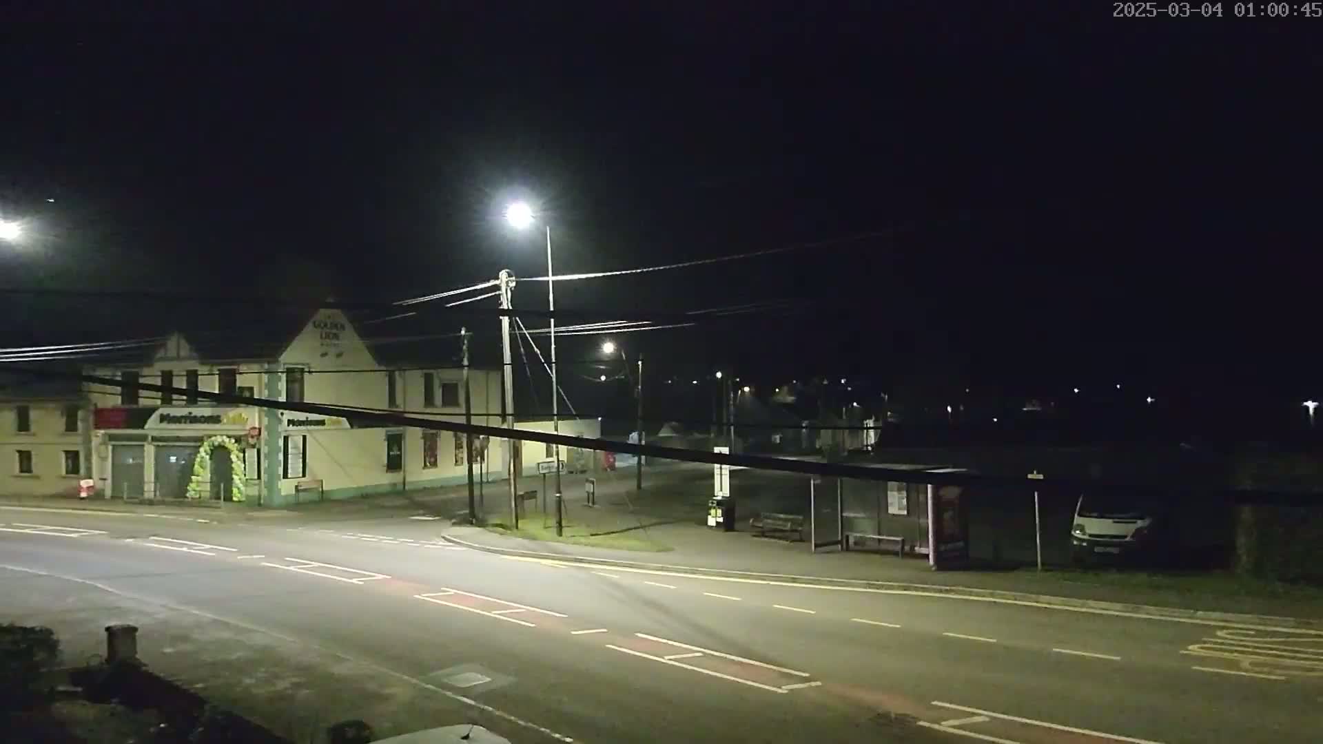 A nighttime street scene shows a crossroads with a building, a bus shelter, and a van parked nearby, under clear skies.