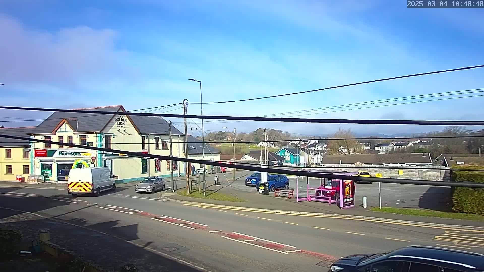 A sunny day reveals a town square with a hotel, a shop, several parked cars, and a bus shelter, overlooked by distant hills.