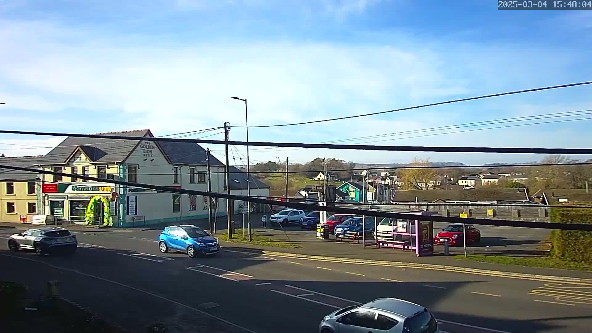 A sunny day reveals a street scene with several cars parked and driving past a two-story building and a small parking lot.