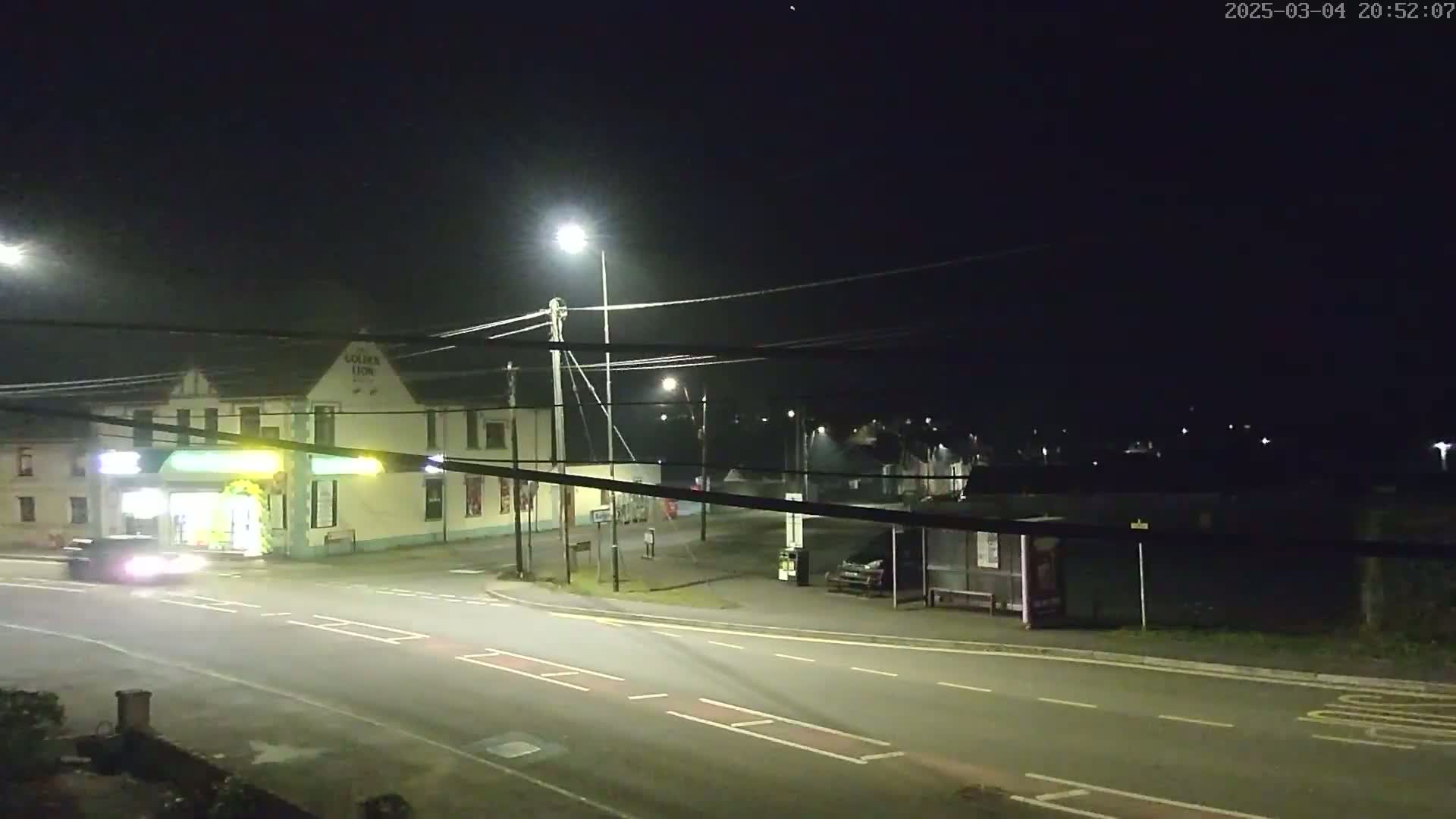 A nighttime street scene shows a car driving past a building with streetlights illuminating a mostly empty road and bus stop.