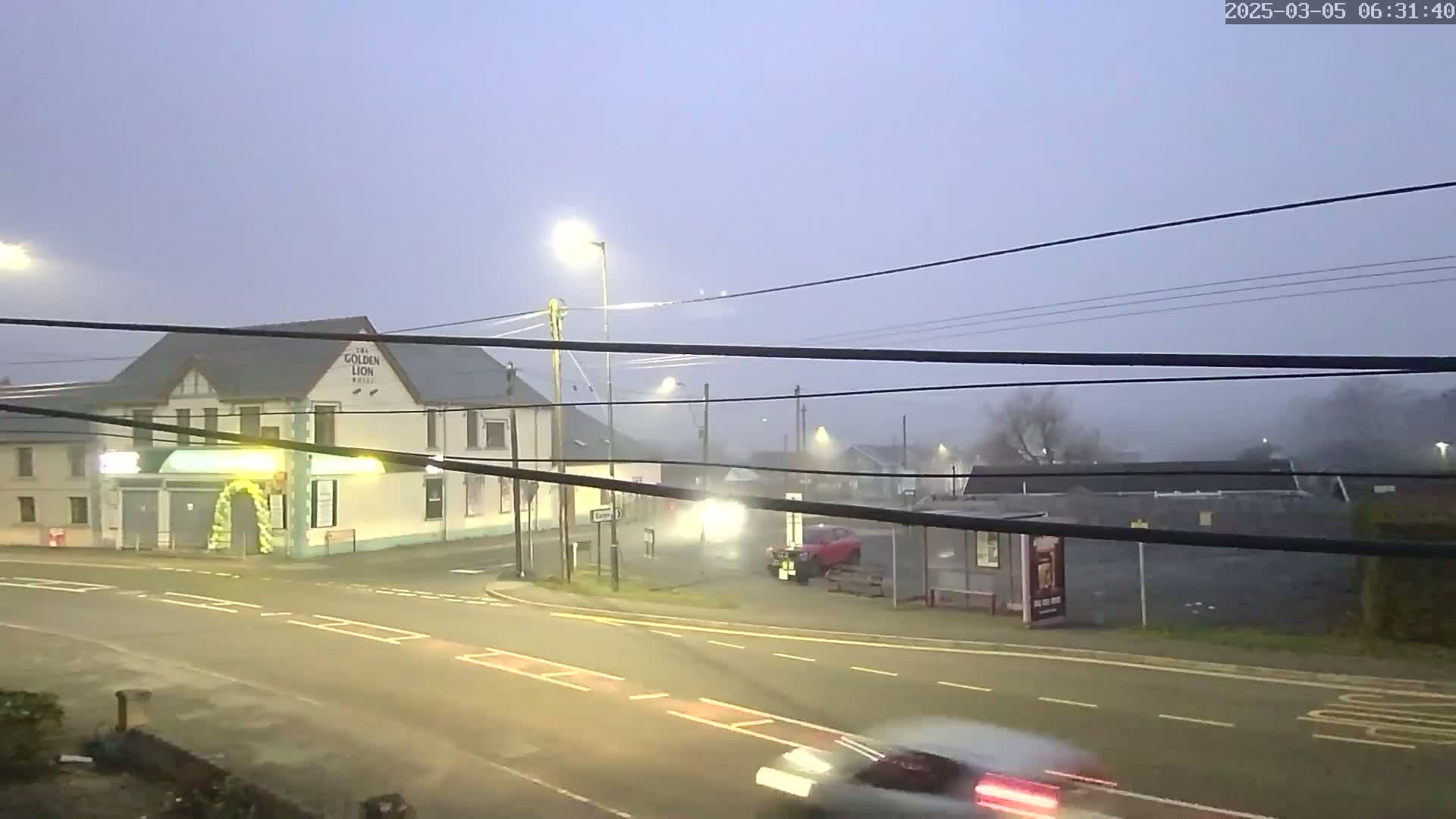 A foggy early morning scene shows a road intersection with a building and several cars, under dim streetlights.