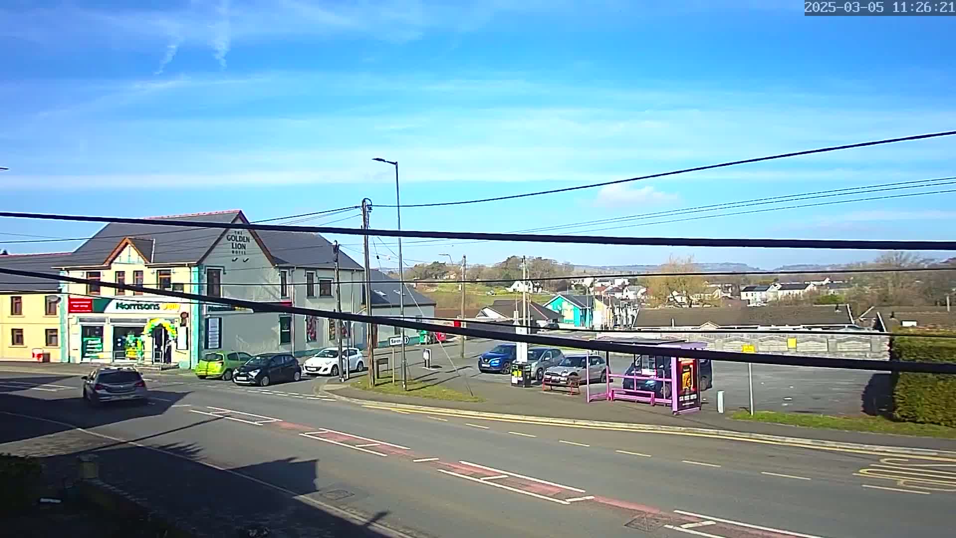 A sunny day reveals a street scene with several cars parked near a bus stop and a two-story building that has a store on the ground floor.