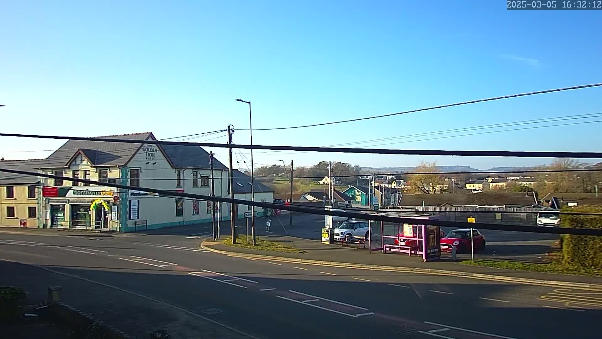 A sunny day reveals a street corner with a hotel, a small shop, cars in a parking lot, and a bus stop, all set against a backdrop of distant hills and houses.