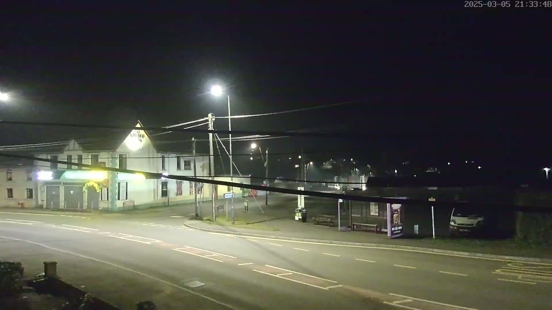 A nighttime street scene shows a building with illuminated signage, a bus stop with a vehicle parked nearby, and a mostly empty roadway under artificial lighting.