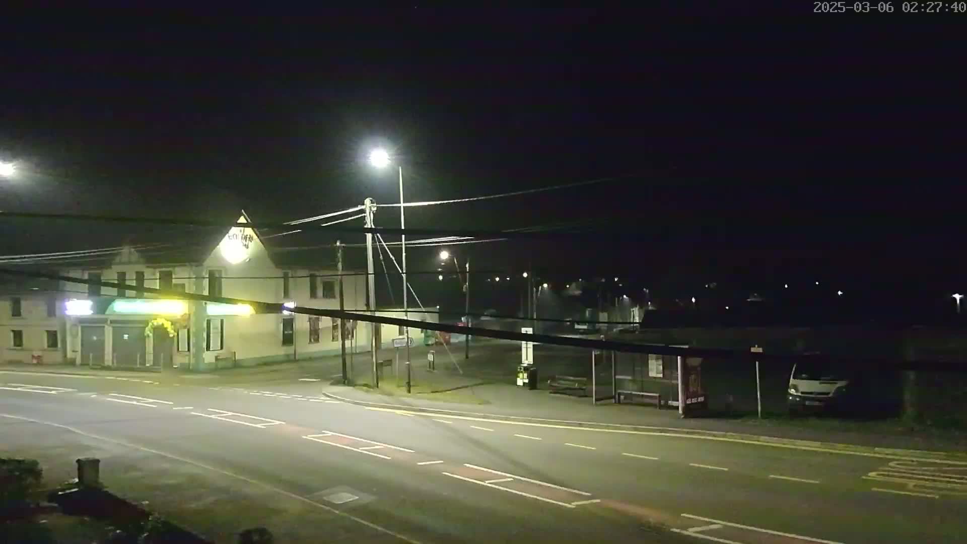 A nighttime street scene shows a two-story building with a lighted sign, a bus stop, and a van parked nearby, illuminated by streetlights under a clear, dark sky.