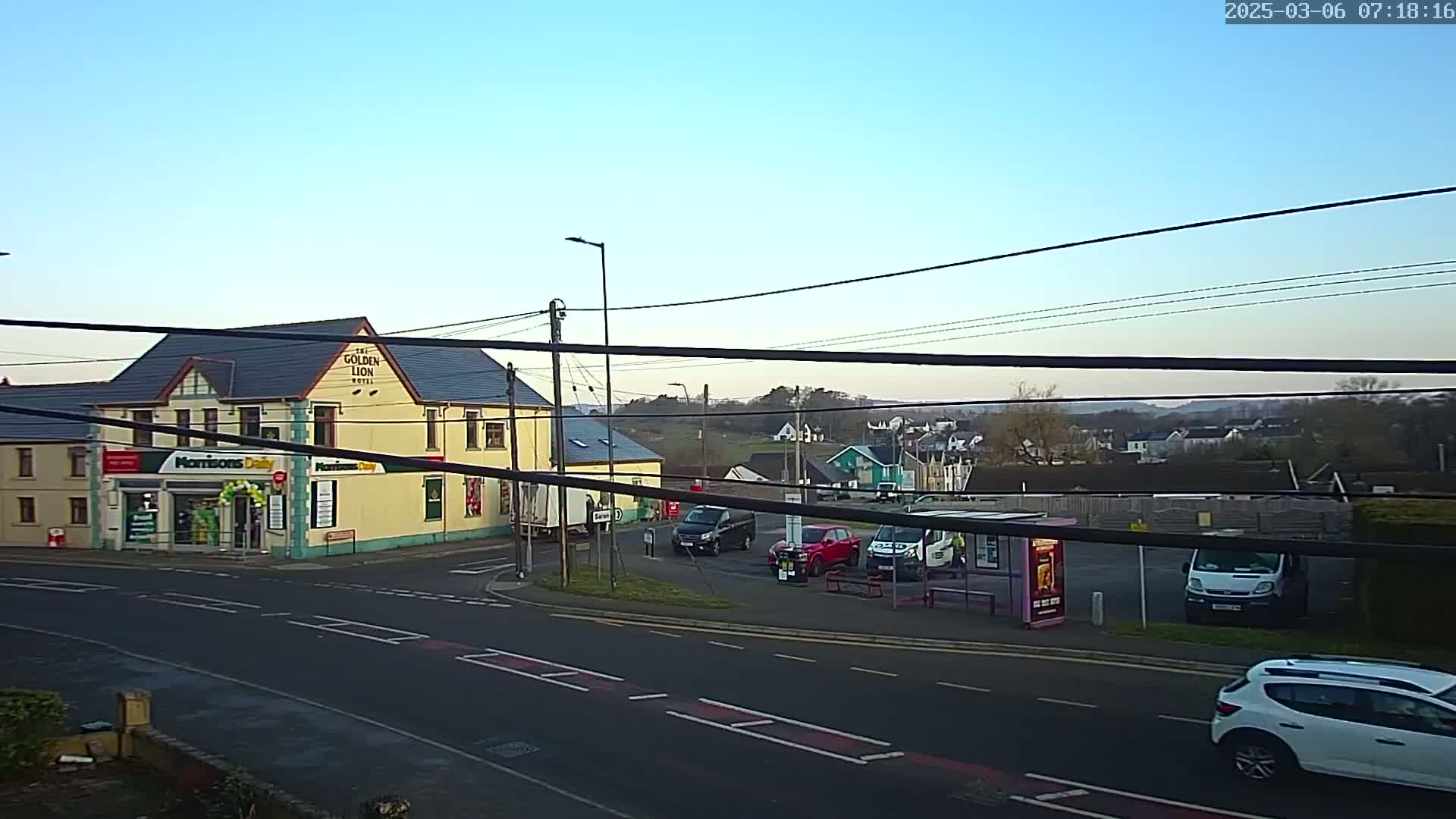 A yellow building with a convenience store and hotel sits at a road intersection under a clear, twilight sky, with several cars parked nearby and a bus stop visible.