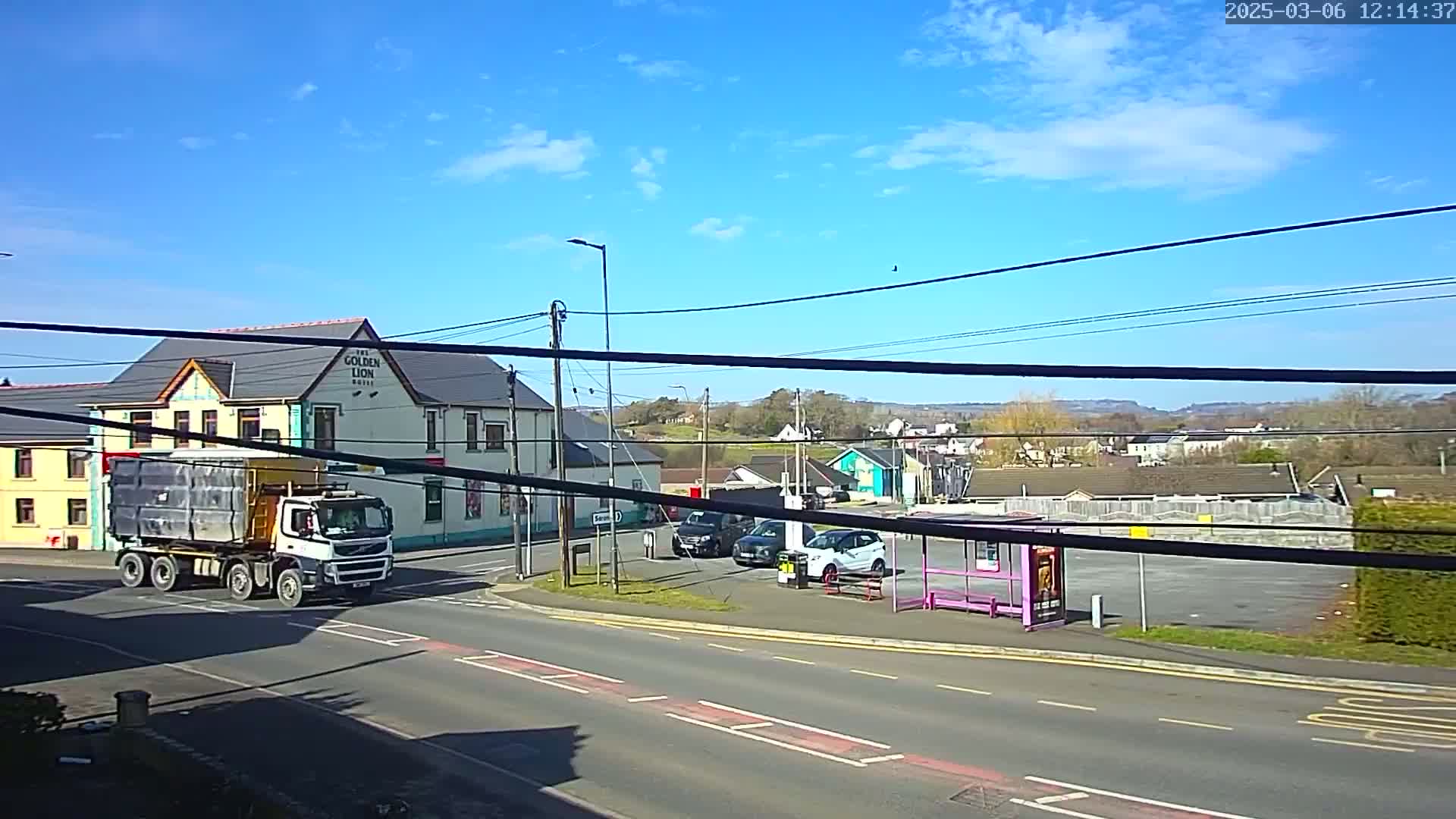 A large truck drives past a pub and a bus stop on a sunny day with few clouds in a town with distant hills.