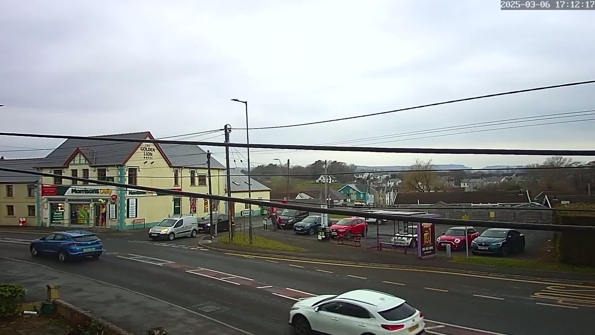 A street scene on an overcast day shows a pub, a small shop, several parked cars, and a few vehicles driving on the road.