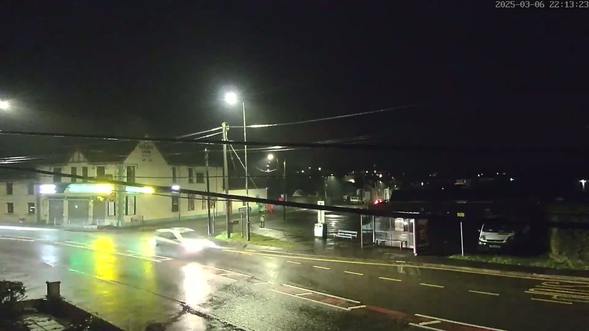 A rain-slicked street at night shows a car driving past a building with lights on, a bus shelter, and a van parked nearby.