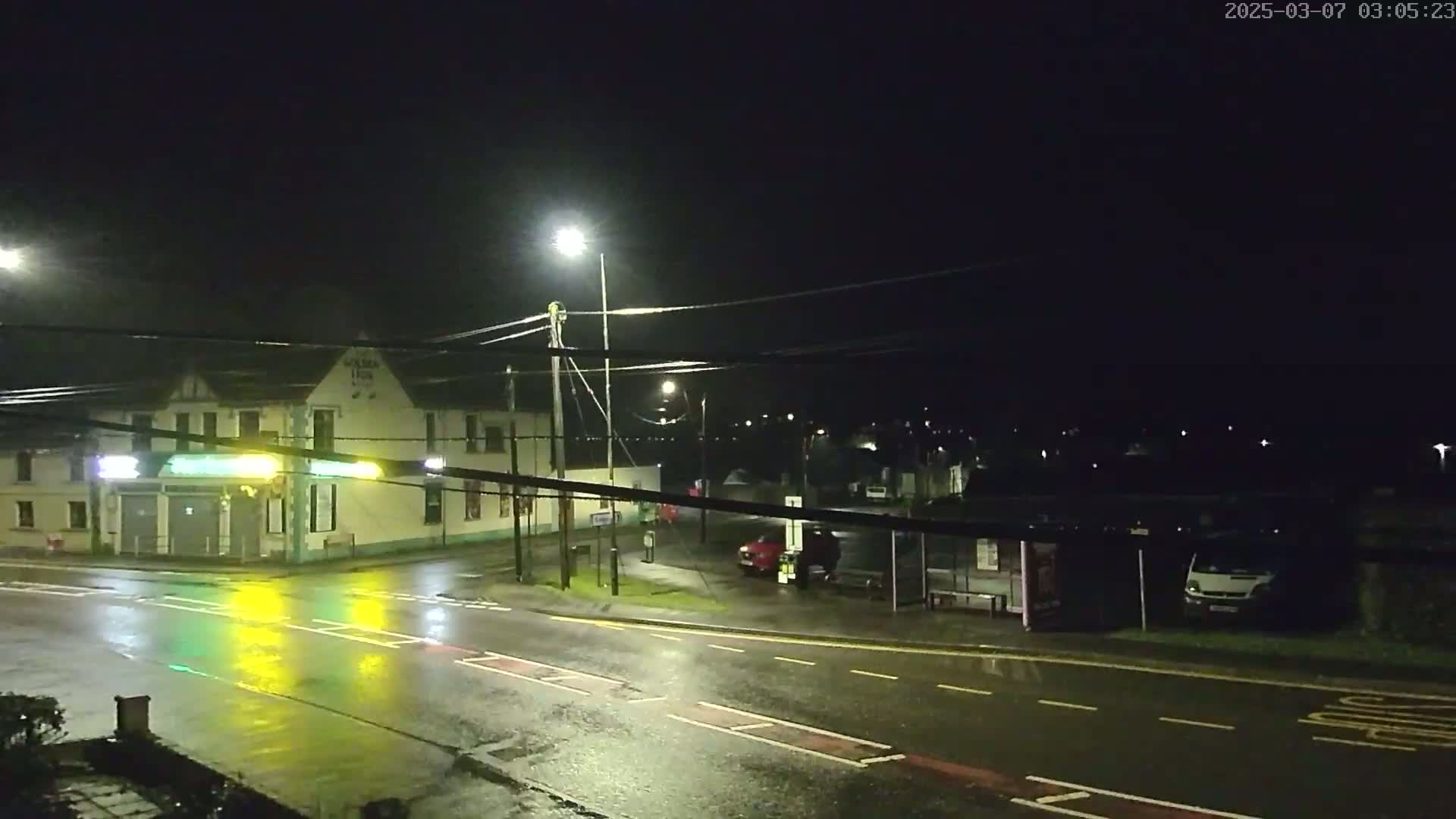 A wet, nighttime street scene shows a building with streetlights illuminating a road, a bus stop with a vehicle, and other vehicles parked nearby.