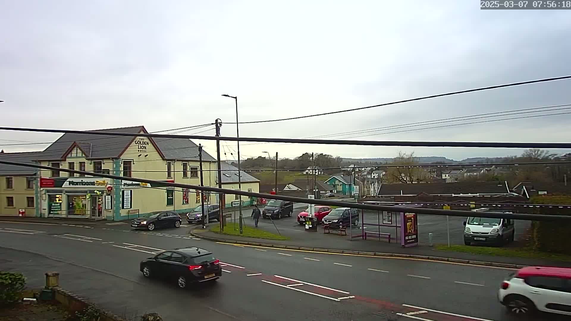 A wet, overcast day reveals a street scene with a pub, a small shop, several cars, and a bus stop, all set against a backdrop of distant hills.