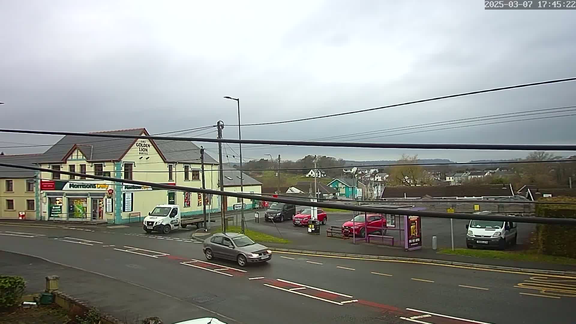 A street scene on an overcast day shows a road with several cars, a bus stop, a small grocery store, and a hotel.