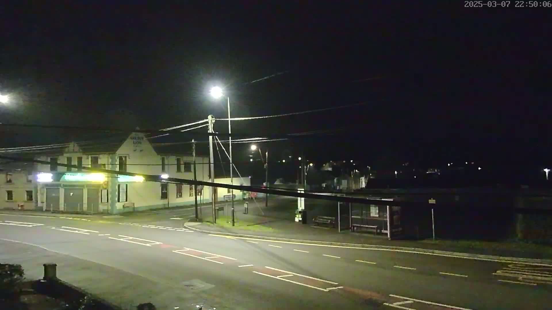 A nighttime street scene shows a corner intersection with a building, streetlights, and a bus shelter under a dark sky.