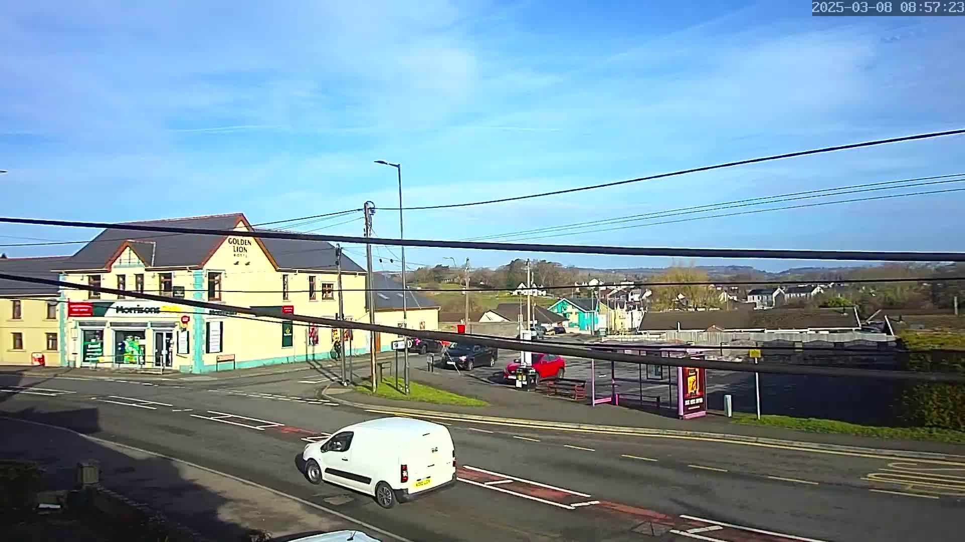 A sunny day reveals a street scene with a yellow building, several cars, a white van, and a bus stop, all situated near a crossroads.